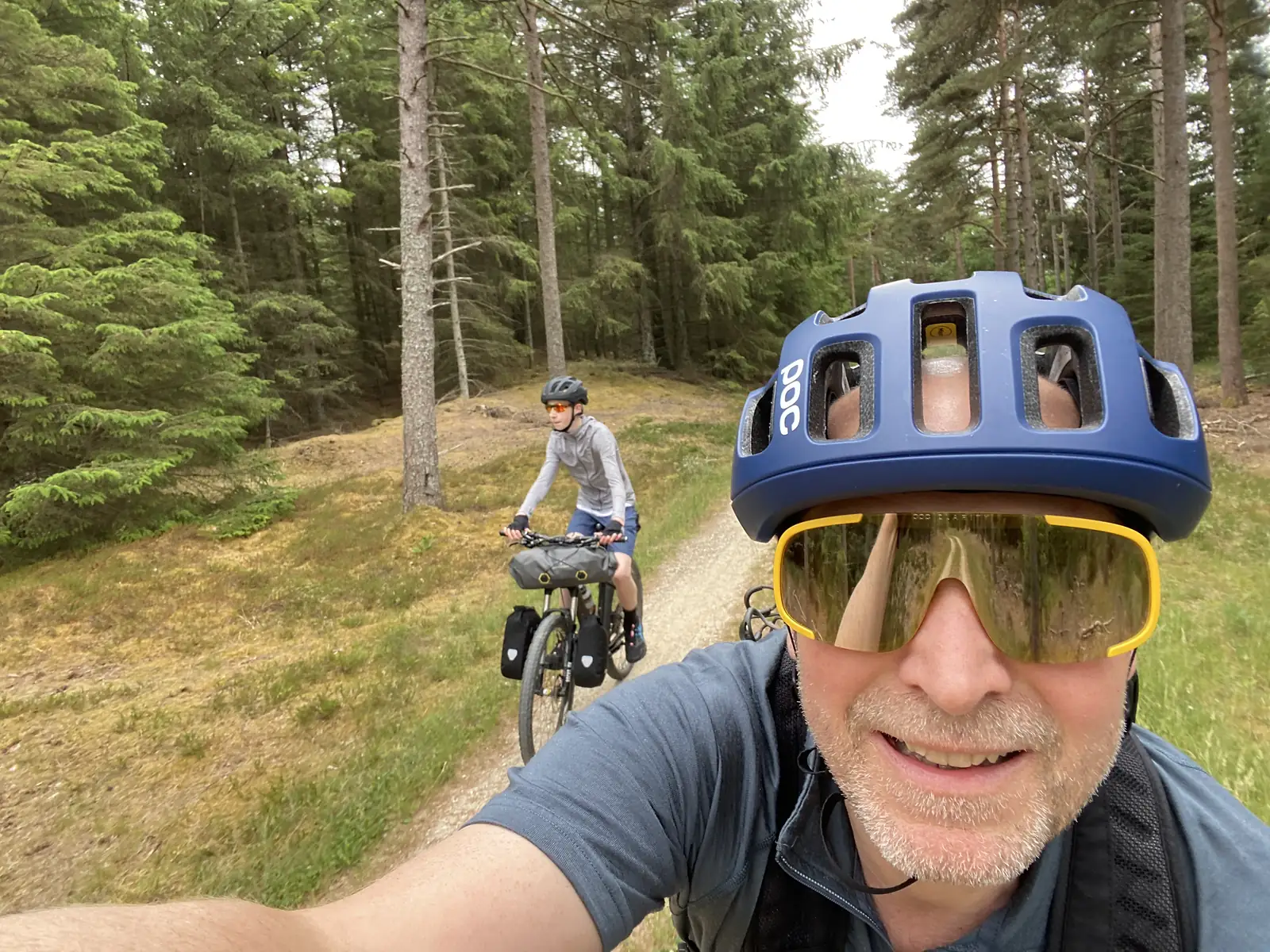 A cyclist in a blue helmet and yellow-rimmed sunglasses grinning close to the camera on a forest track, with a second rider on a loaded bike coming up behind.