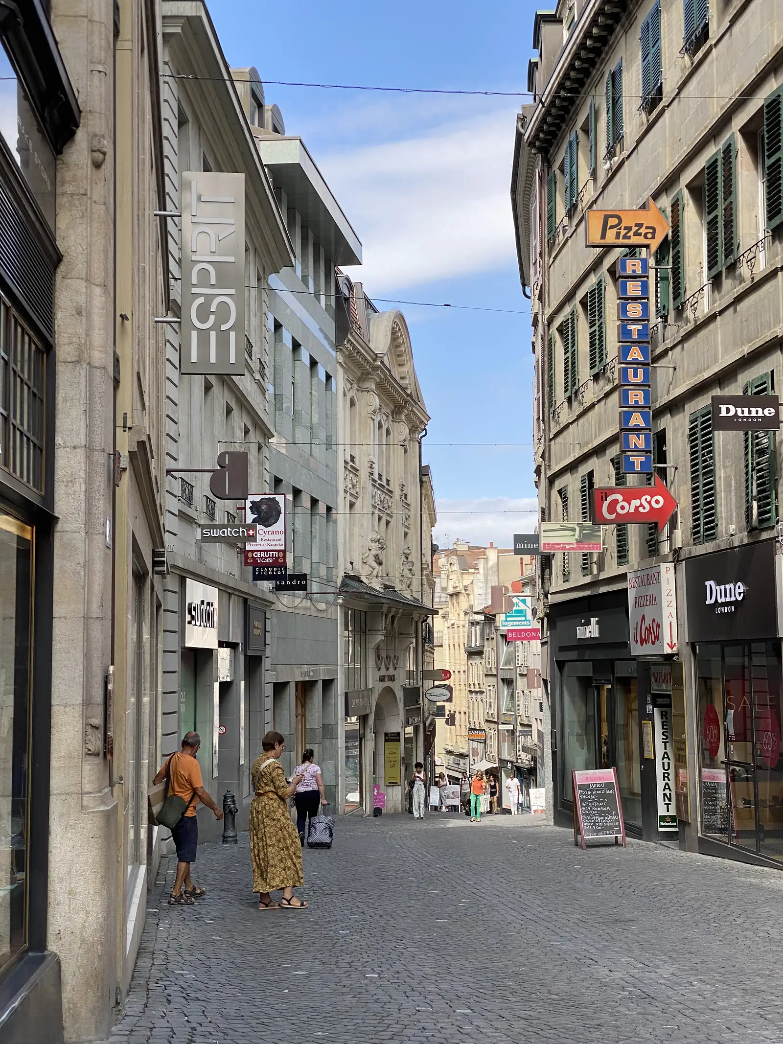 A narrow Lausanne shopping street hung with signs for Esprit, Swatch and an Italian restaurant advertising Pizza and il Corso, pedestrians strolling the cobbles below.