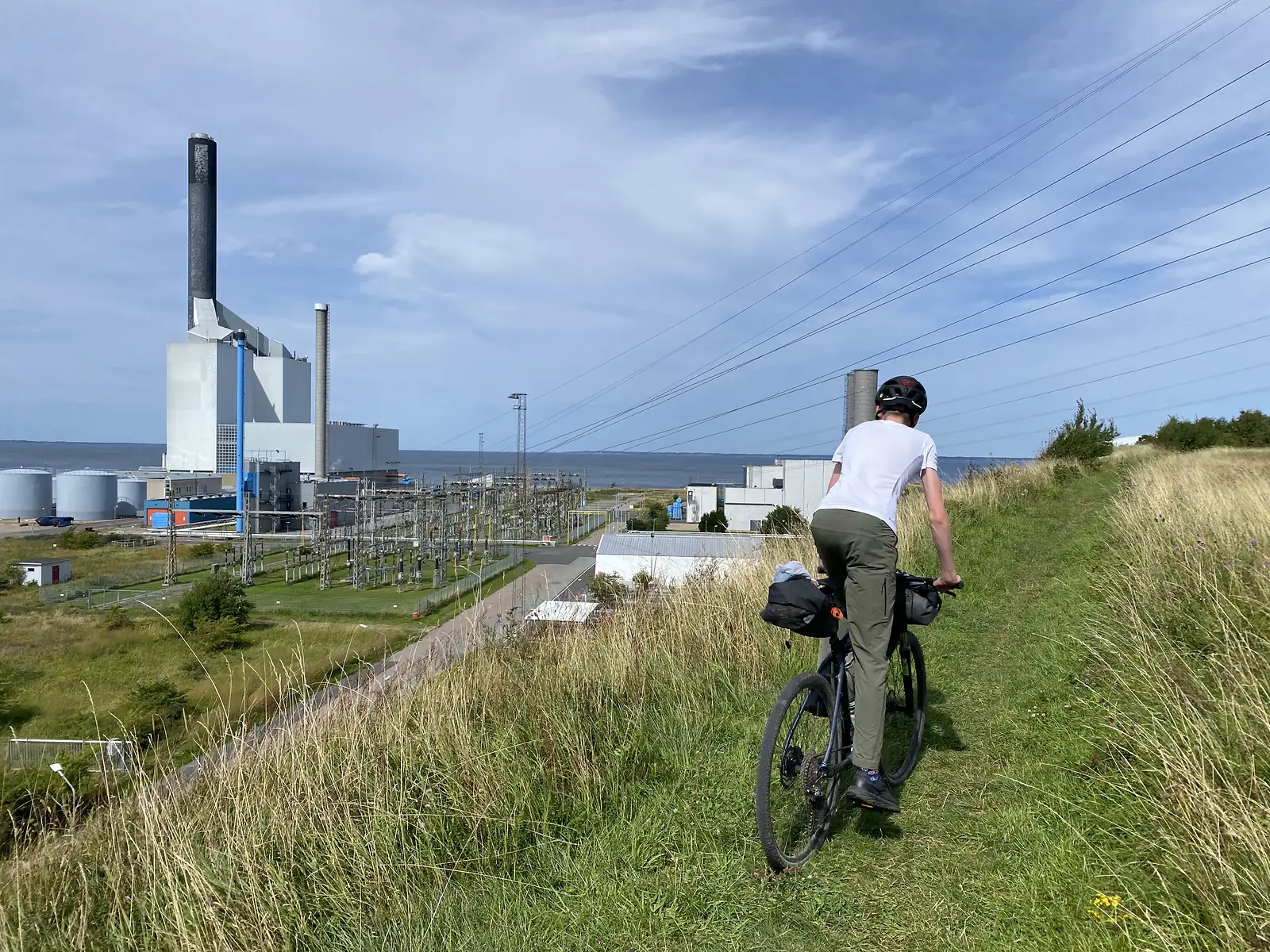 A rider on a grassy path above Kyndbyværket power station on Isefjord, its chimney, storage tanks and switchyard below.