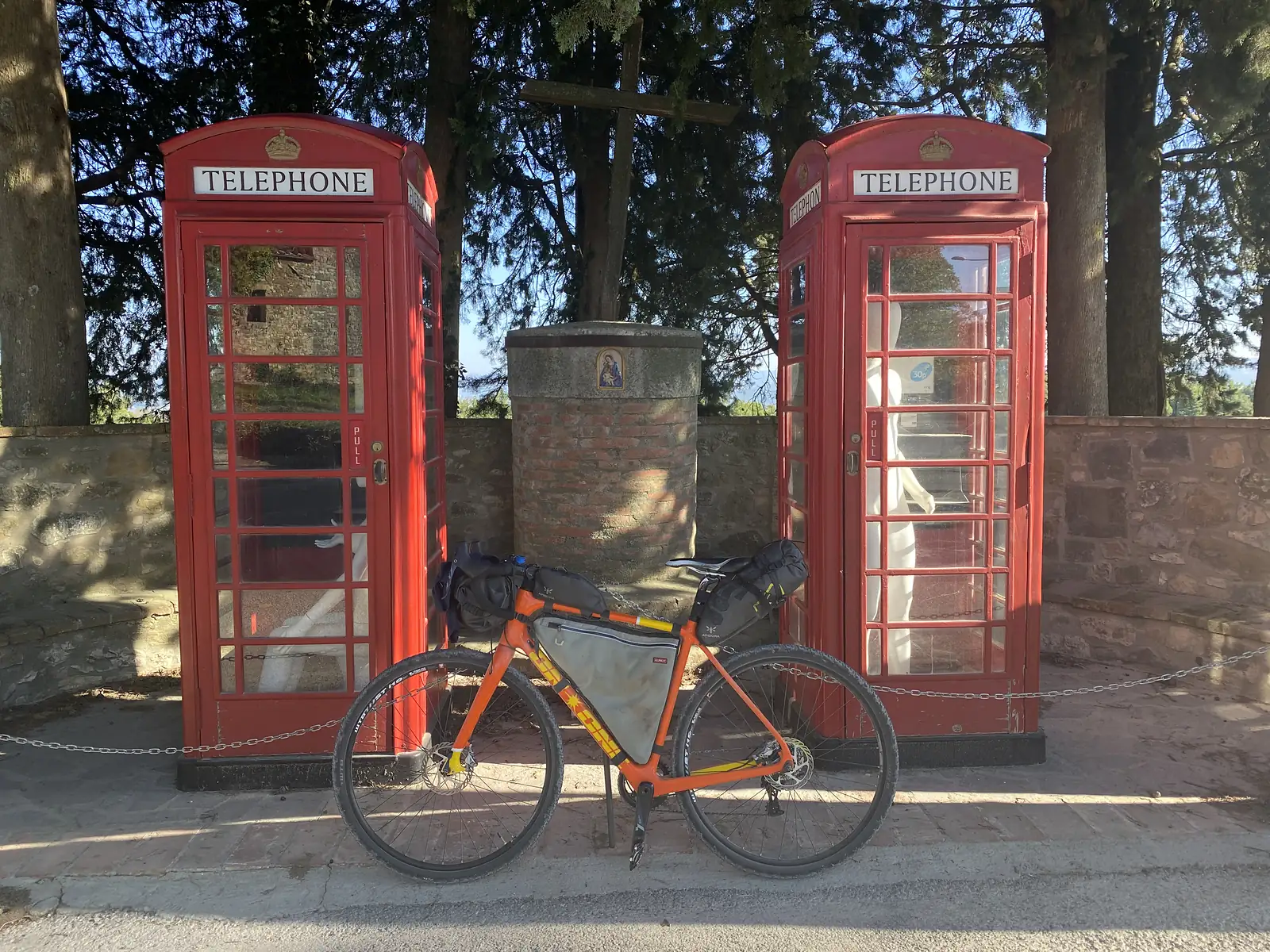 A loaded orange bikepacking bike parked in front of two red British-style telephone boxes, each with a mannequin inside, flanking a small brick shrine.