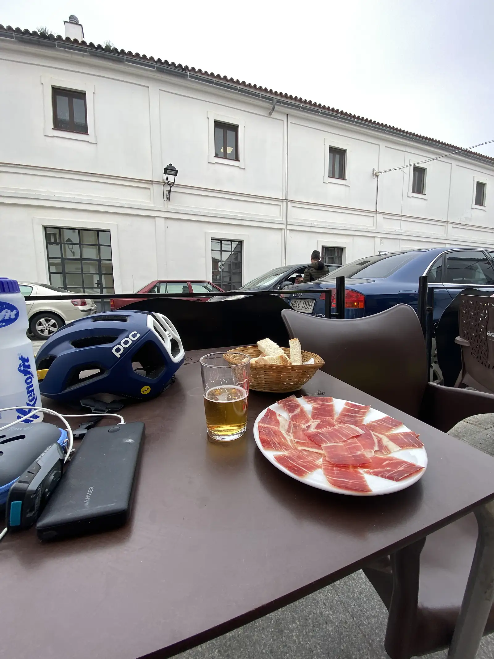 A plate of thinly sliced cured ham, a glass of beer and a basket of bread on a pavement table, with a blue cycling helmet set down beside them.