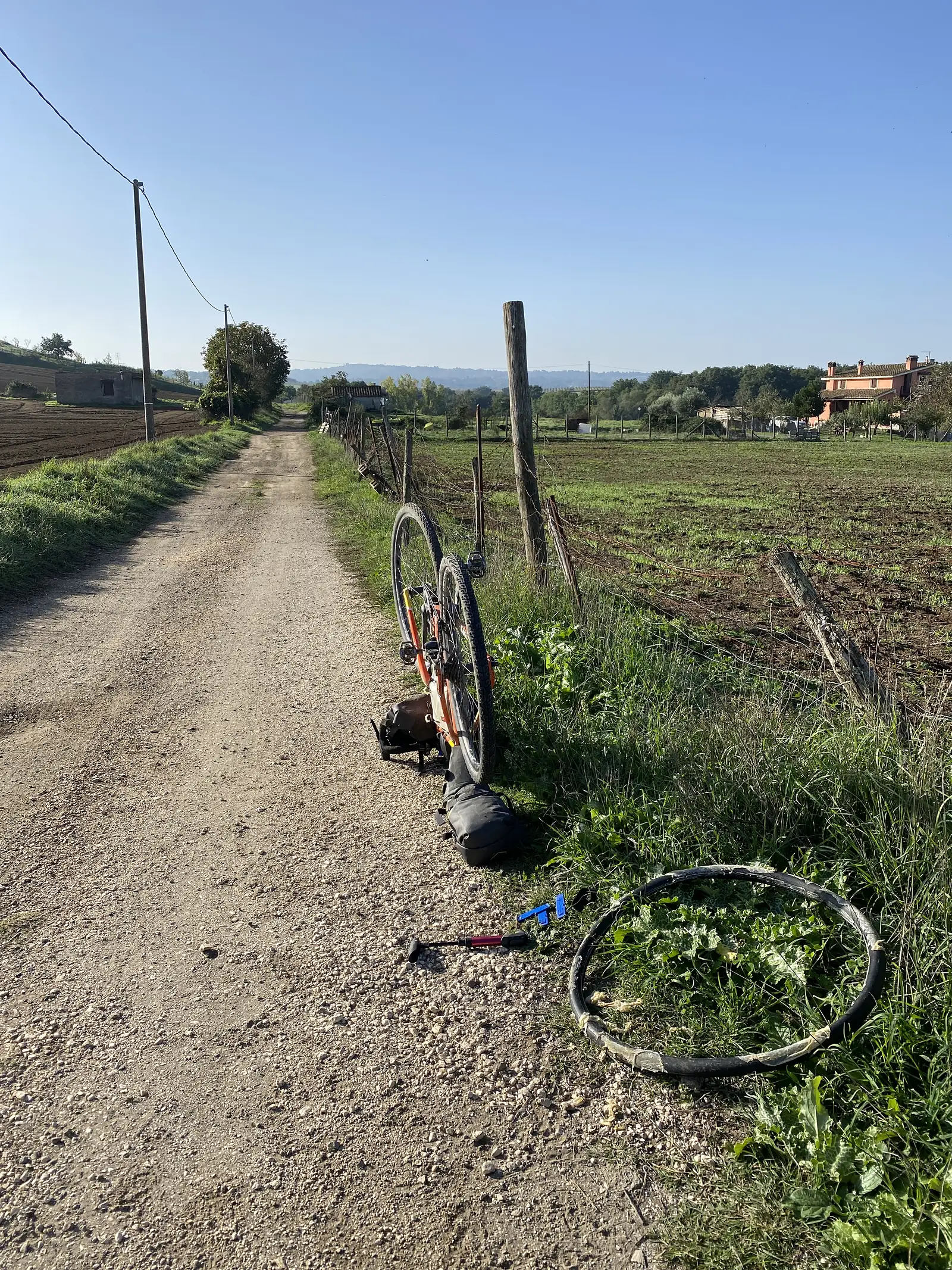 A mid-repair puncture stop beside a gravel track, with the bicycle's tyre and rim removed, a mini pump, and a tyre lever laid out on the ground.