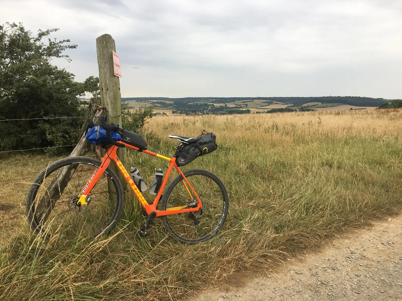 An orange gravel bike loaded with bikepacking bags leans against a wooden waymarker post beside a barbed-wire fence, overlooking rolling arable fields and a distant village.