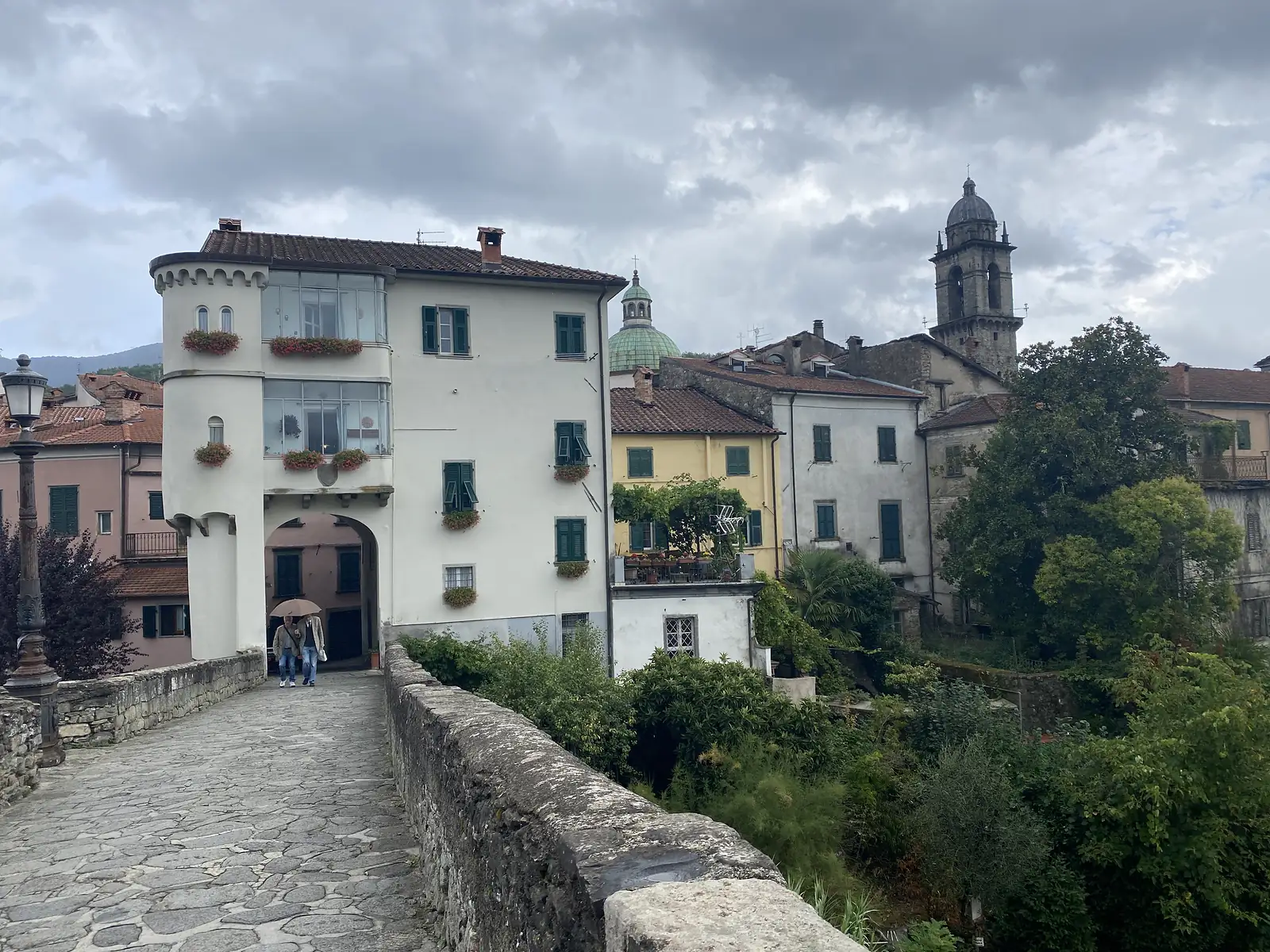 Two people with an umbrella walk beneath a fortified gate tower guarding a stone bridge into an old town, church domes and a bell tower rising above the rooftops beyond.