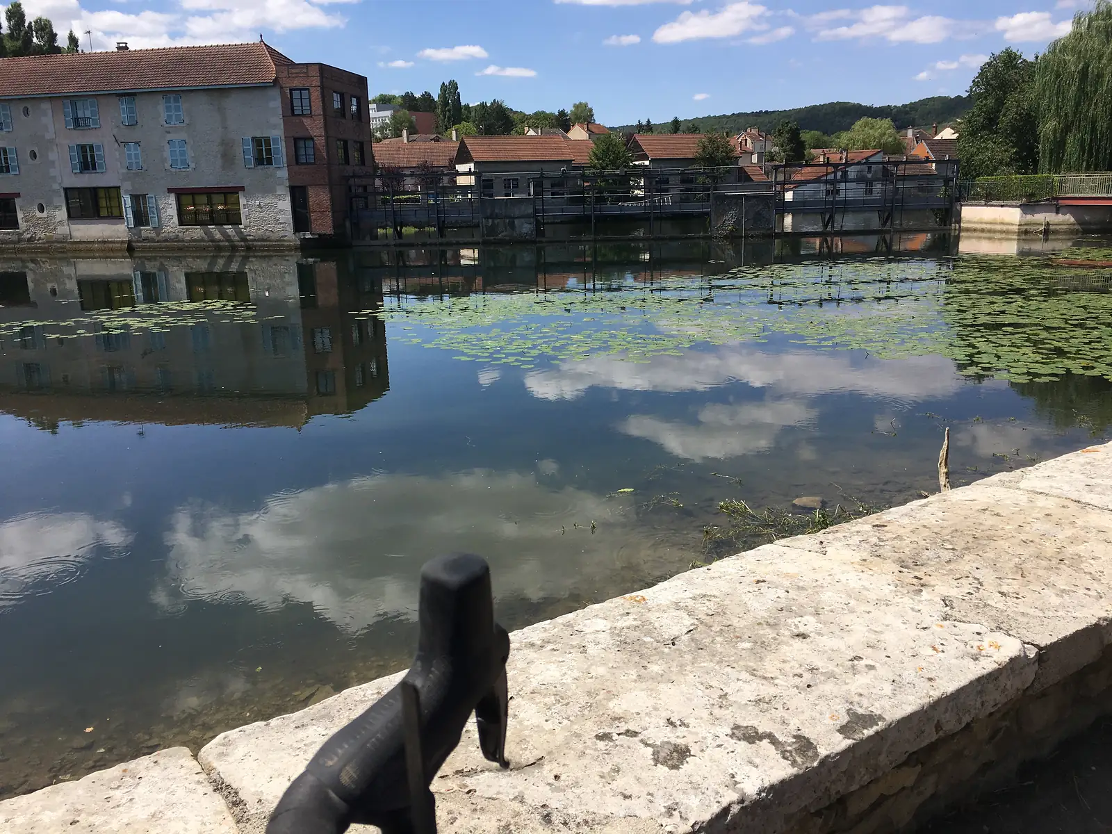 A lily-covered stretch of river runs past stone houses with pale blue shutters, with a weir and metal walkway spanning the water.