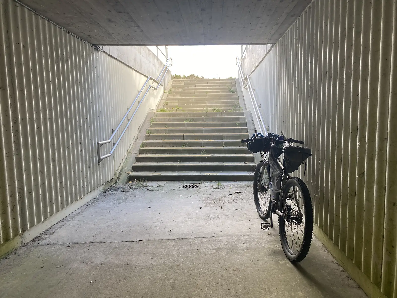 A loaded bikepacking bike standing in a concrete underpass at the foot of a long flight of steps leading up to daylight.