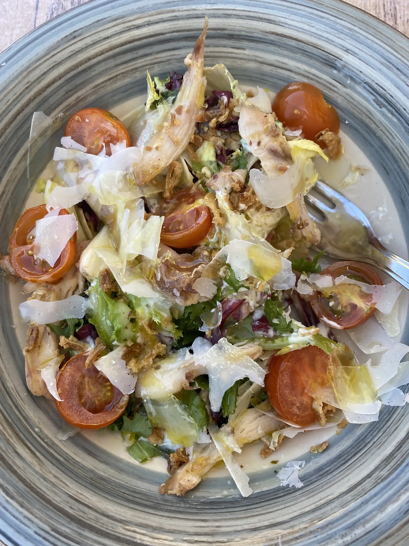 A bowl of chicken Caesar salad with cherry tomatoes, shaved cheese, crispy onions and dressed leaves.