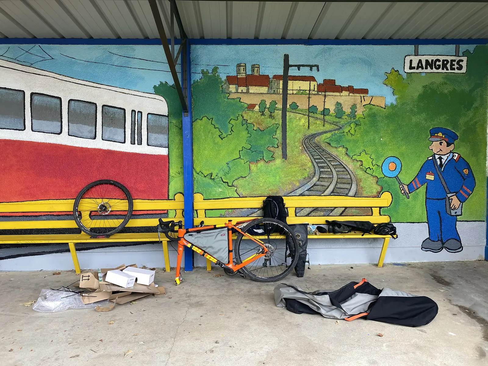 A loaded orange gravel bike leans against a yellow bench painted with a mural of a train and a signalman beside a sign reading 'LANGRES', with packaging cardboard strewn on the ground where the bike has just been unboxed.