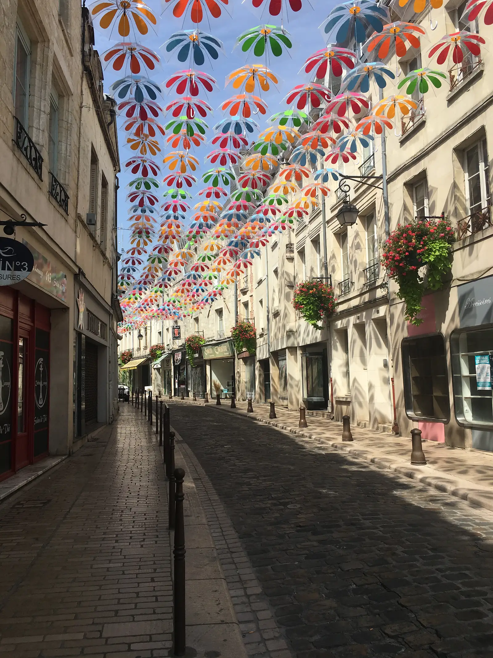A canopy of colourful patterned umbrellas stretches overhead along a street of pale stone shopfronts hung with baskets of red flowers.
