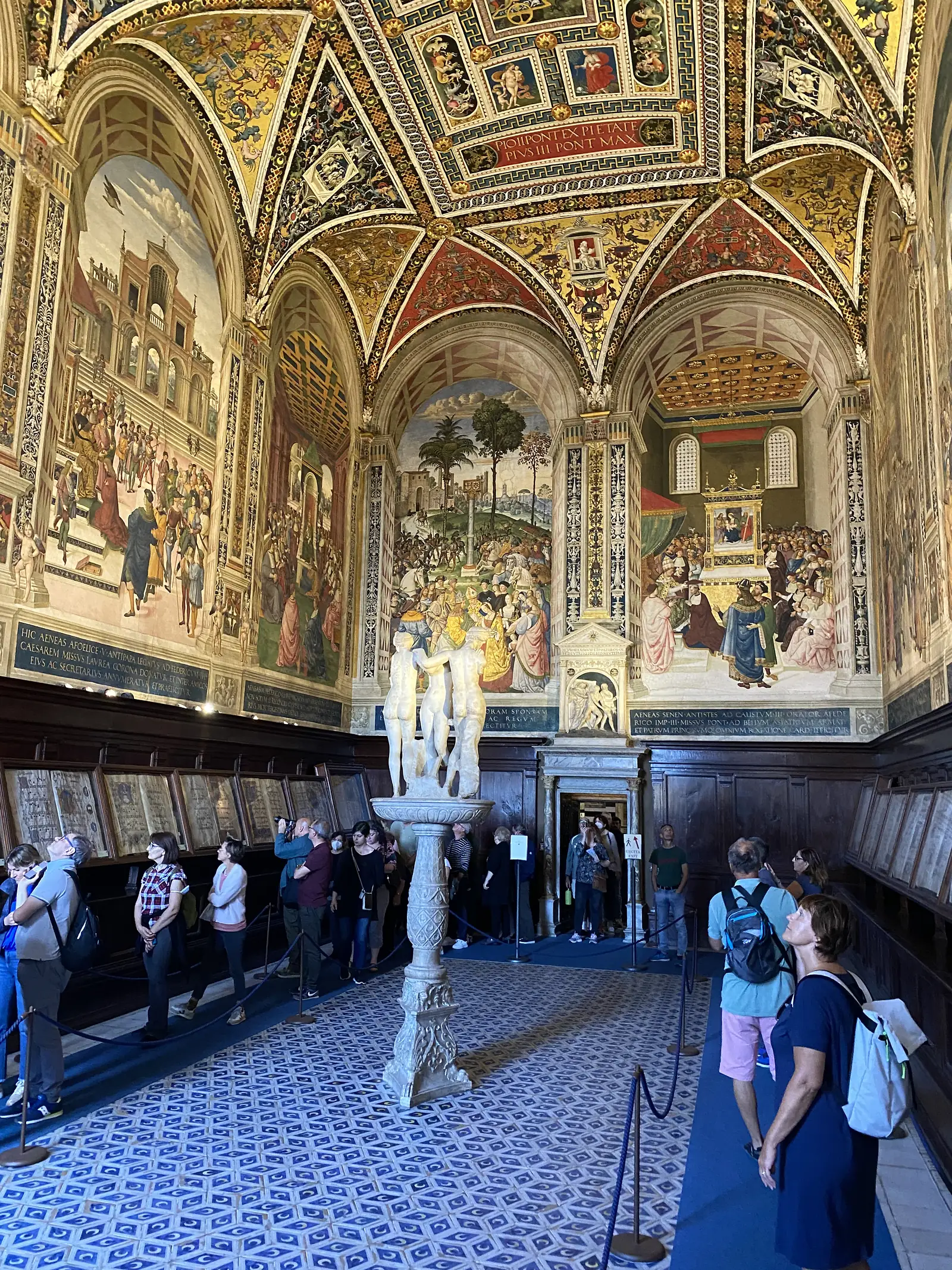 Visitors gathered beneath the vividly frescoed vaulted ceiling of a Renaissance library, with a marble statue on a pedestal in the centre of the room.
