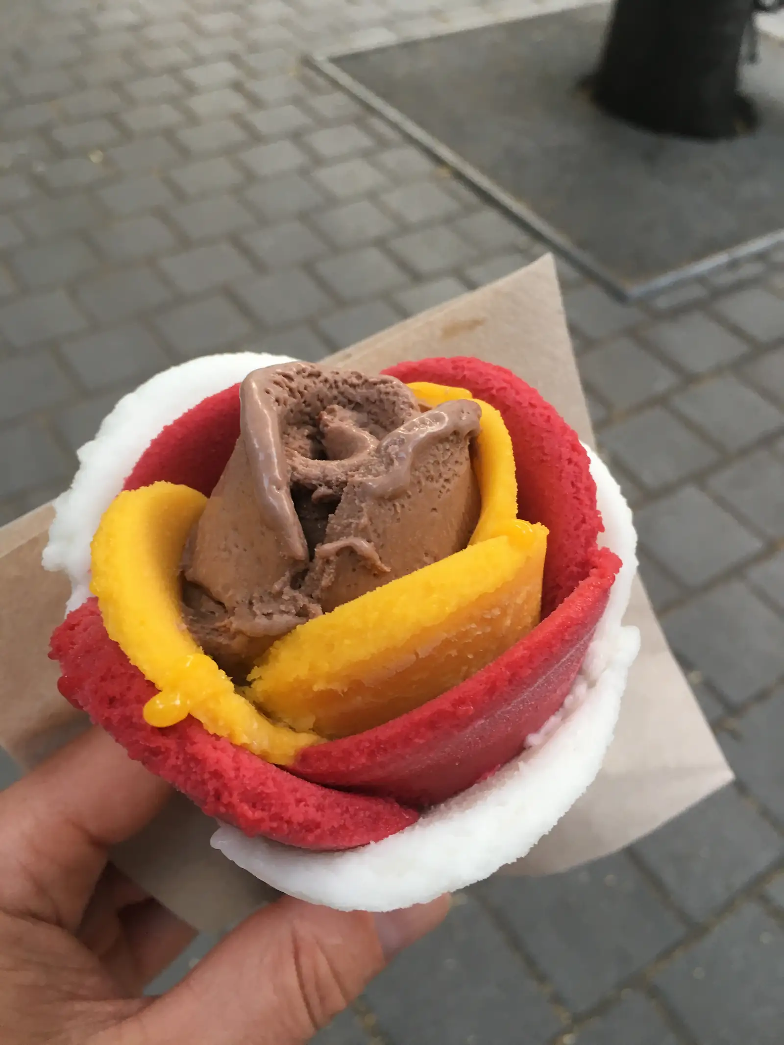 A hand holds a scoop of chocolate sorbet nestled between wafers of red, yellow and white flavoured biscuit, dripping onto a paper wrapper.