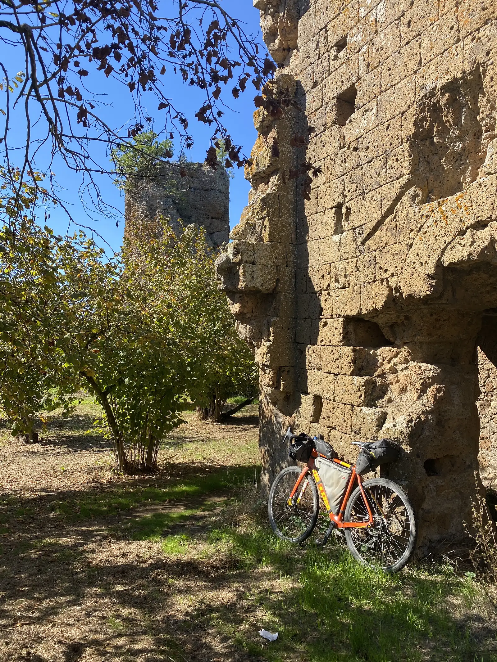 An orange loaded touring bike with panniers leaning against the weathered brick pillars of an ancient ruined arch, more remains visible in the field beyond.