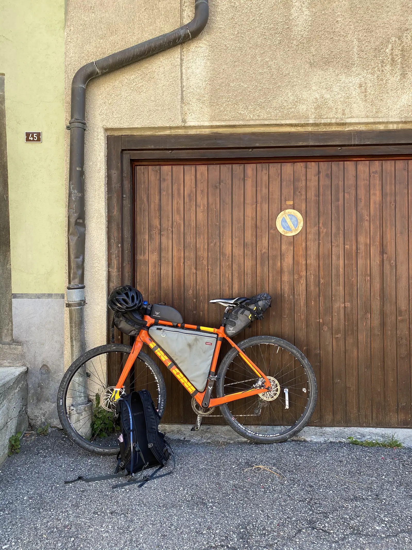 A loaded orange touring bike with a helmet and rucksack propped against a wooden garage door beside house number 45.