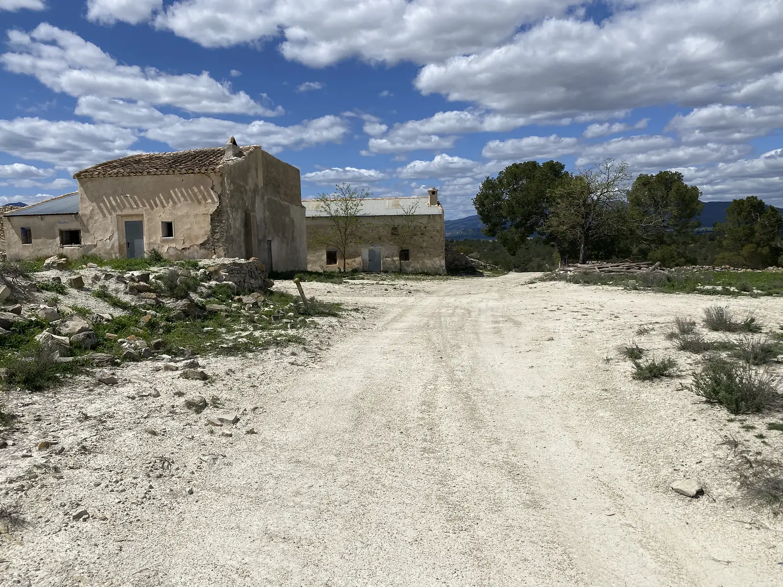 An abandoned farmstead of rendered stone buildings with a tiled roof and corrugated outbuildings, standing beside a bare white track.