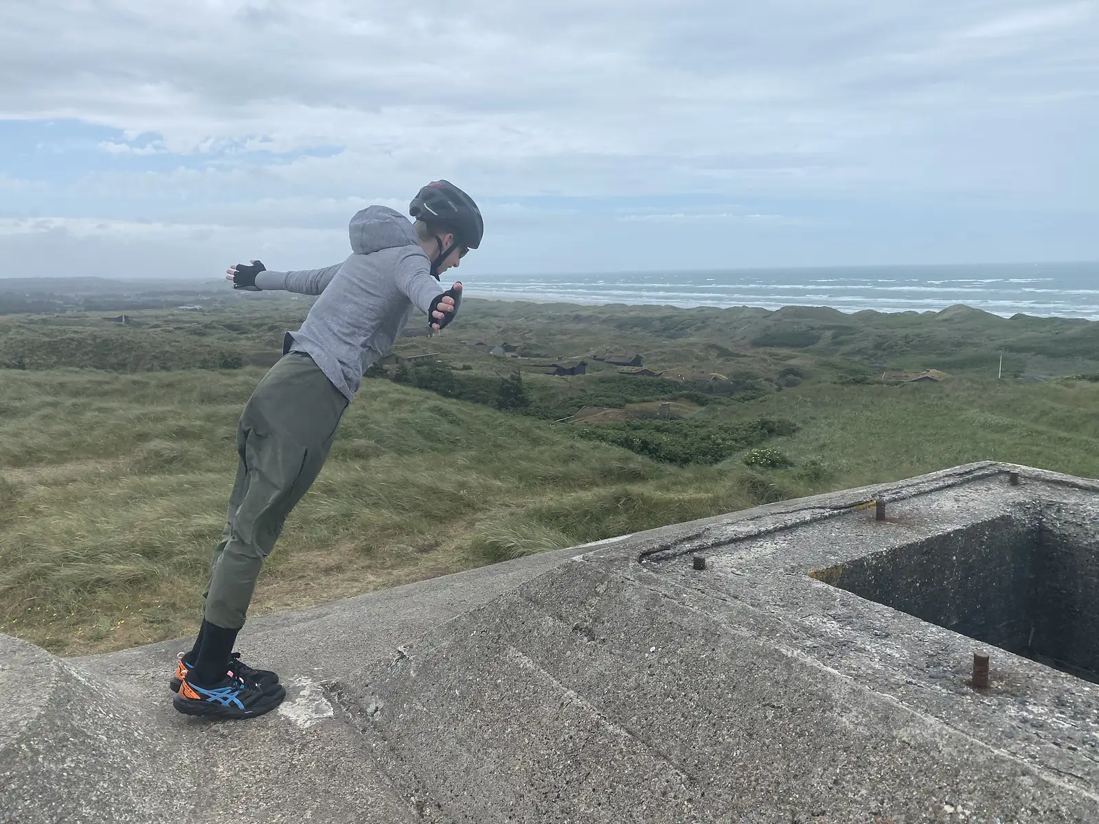 A young person in a cycling helmet leaning into the wind with arms outstretched on top of a concrete bunker above the dunes and sea.
