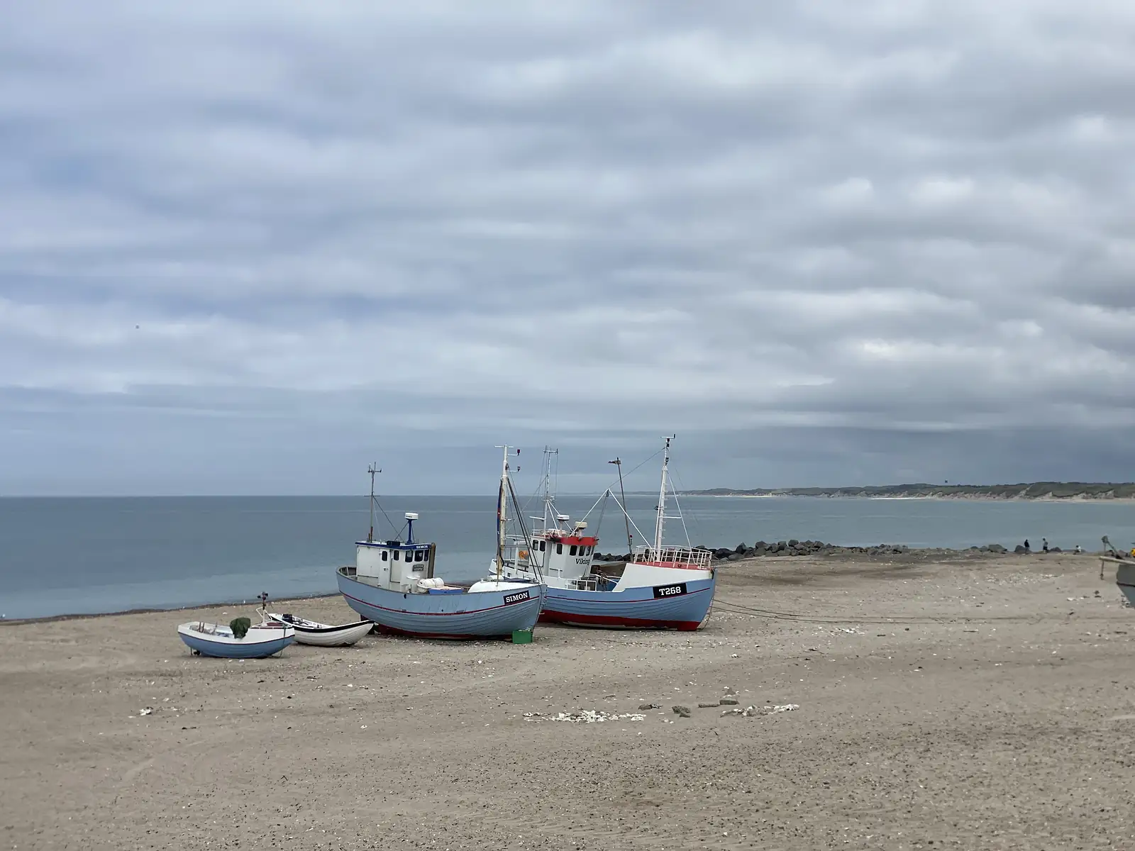 Two clinker-built fishing boats hauled up on a sandy beach, one lettered "SIMON", with dinghies alongside and a rock breakwater beyond.