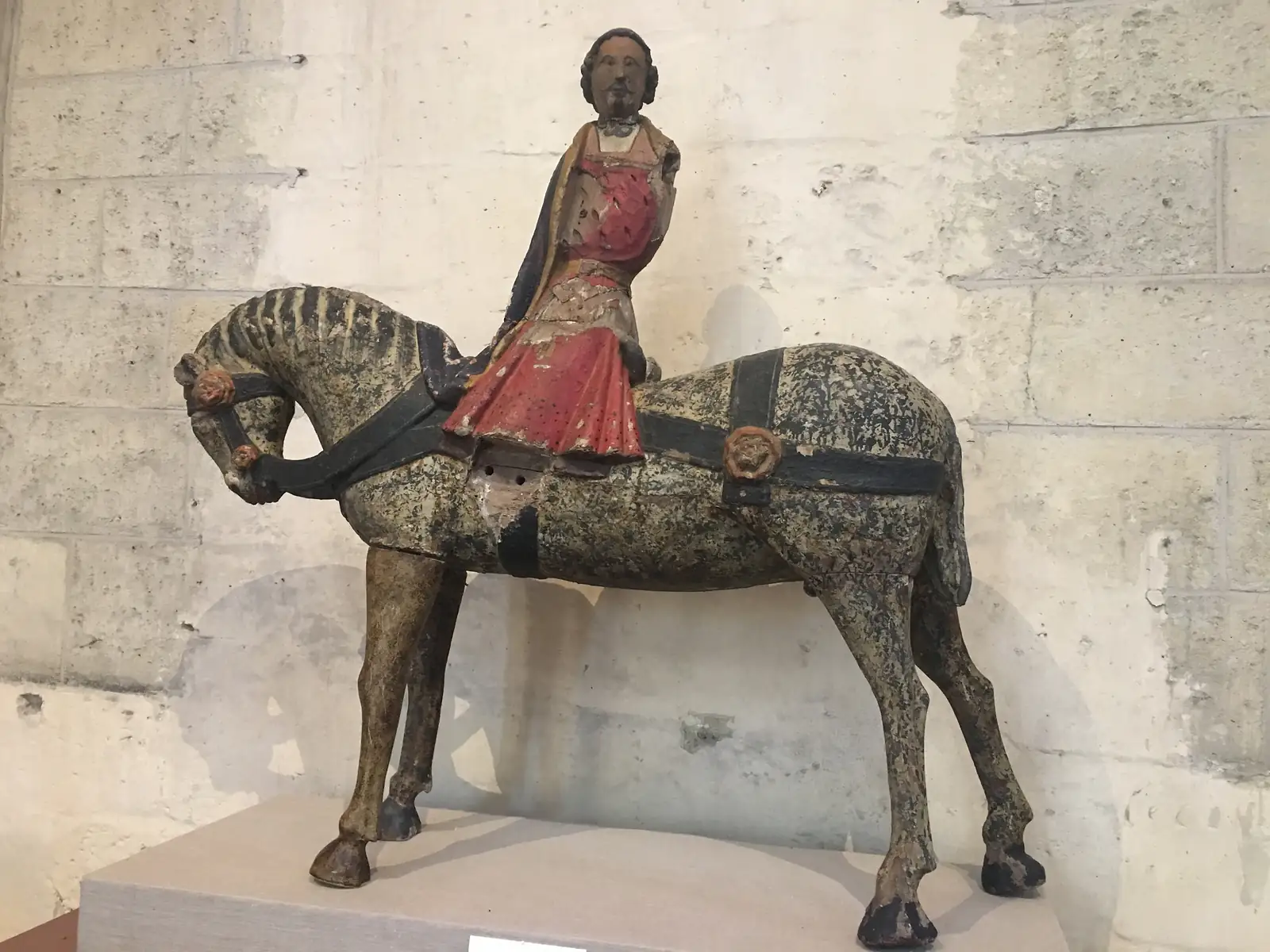 A weathered polychrome wooden statue of a robed figure riding a horse stands on display against a bare stone wall.
