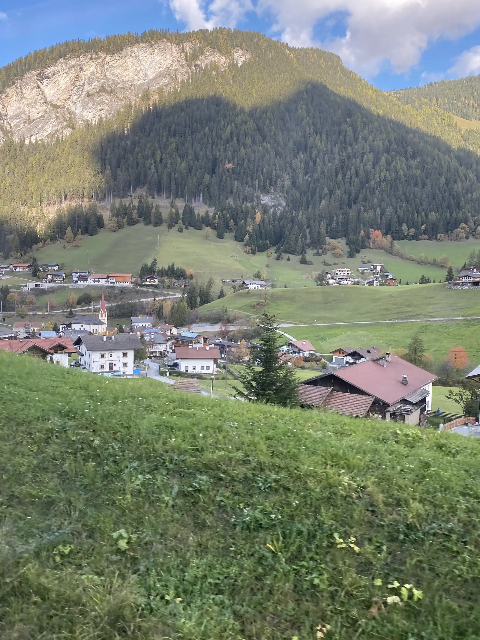 An alpine village with a church spire sits in a green valley beneath forested slopes and a bare limestone cliff.