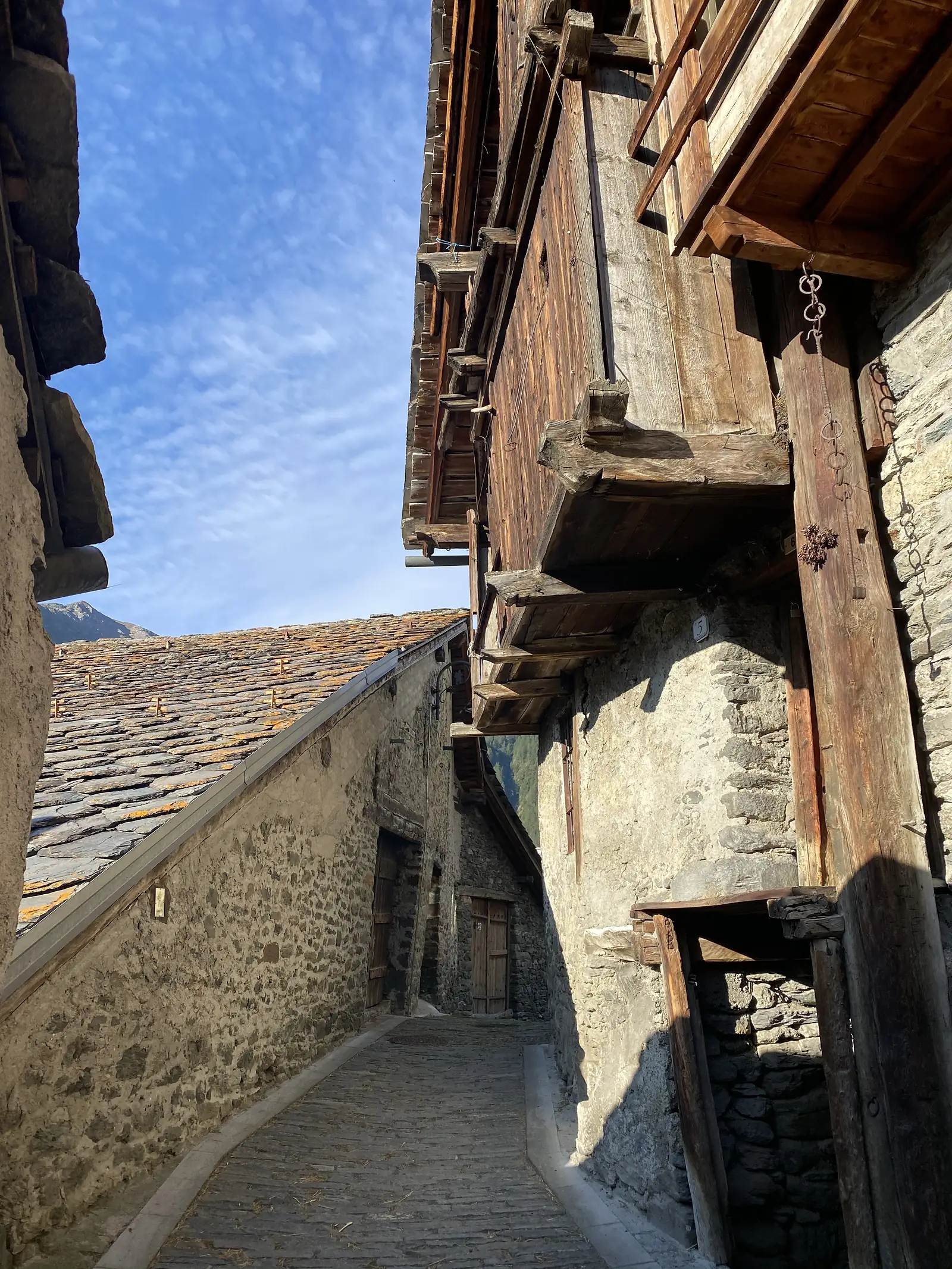A narrow cobbled alley winds between old stone houses with jutting timber balconies and stacked slate roofs, sunlight breaking through at the far end.