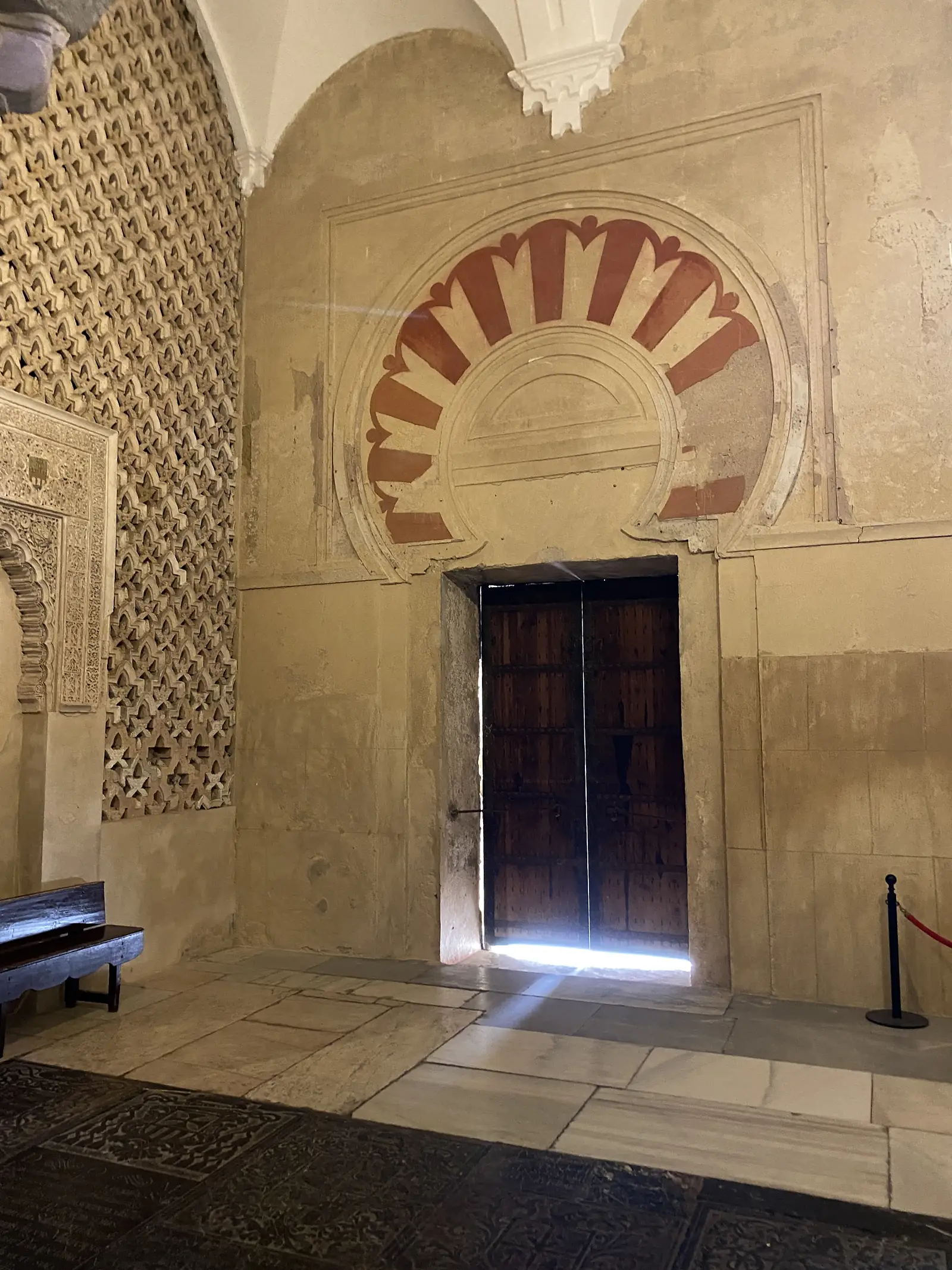 A studded wooden door set beneath a red-and-white scalloped horseshoe arch, beside a wall panelled with intricate interlaced stonework.
