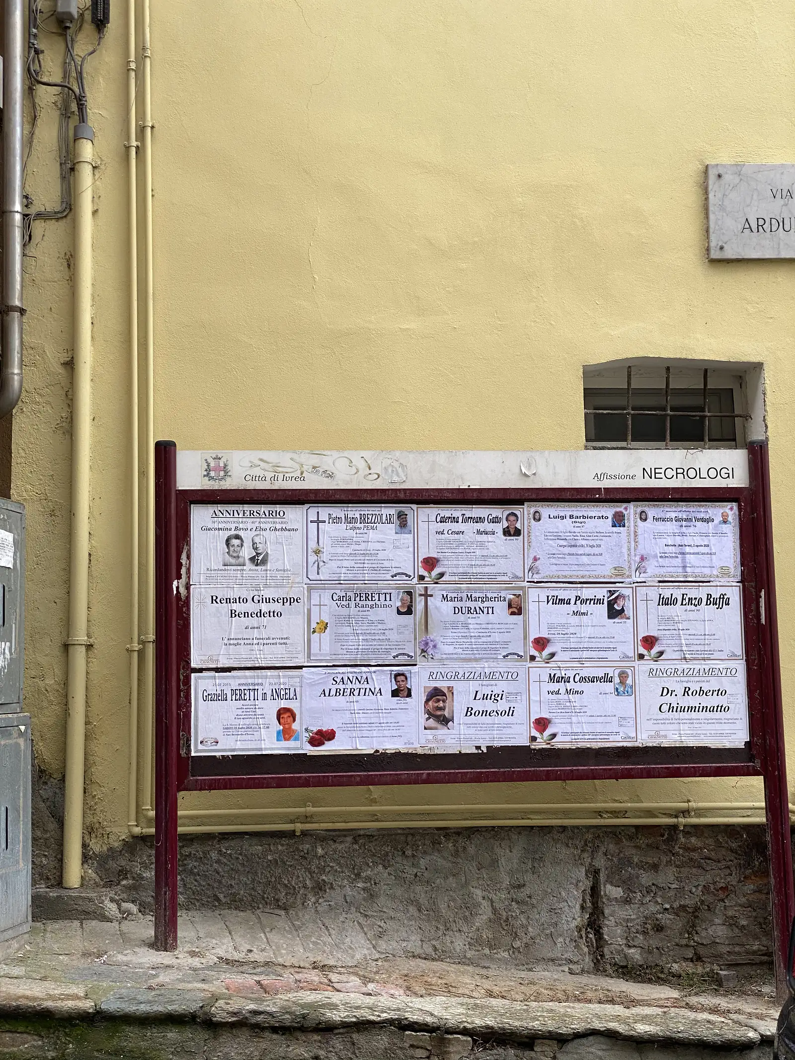 A public noticeboard headed 'Città di Ivrea, Affissione Necrologi' mounted on a yellow wall is covered with local death notices and memorial cards.