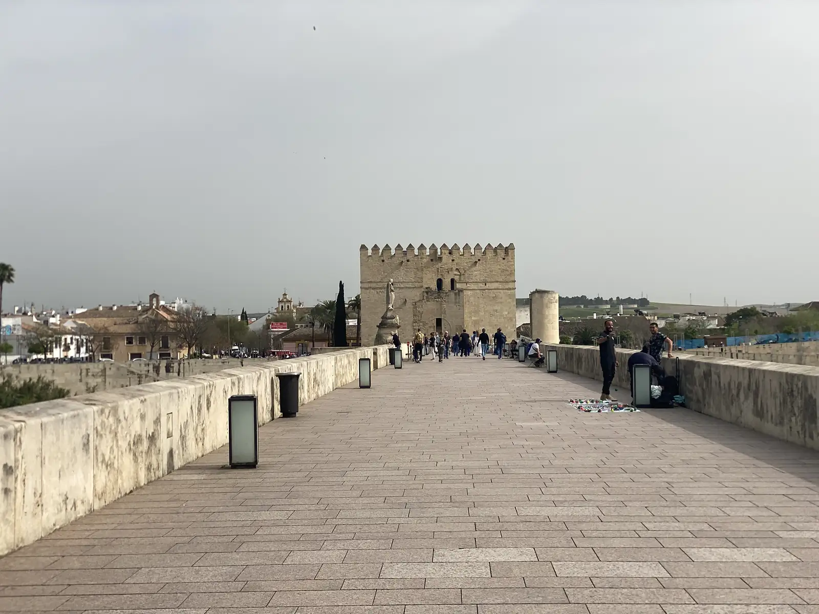 A wide paved bridge walkway leading towards a battlemented stone gate tower, with strollers and a street trader's wares laid out on the ground.