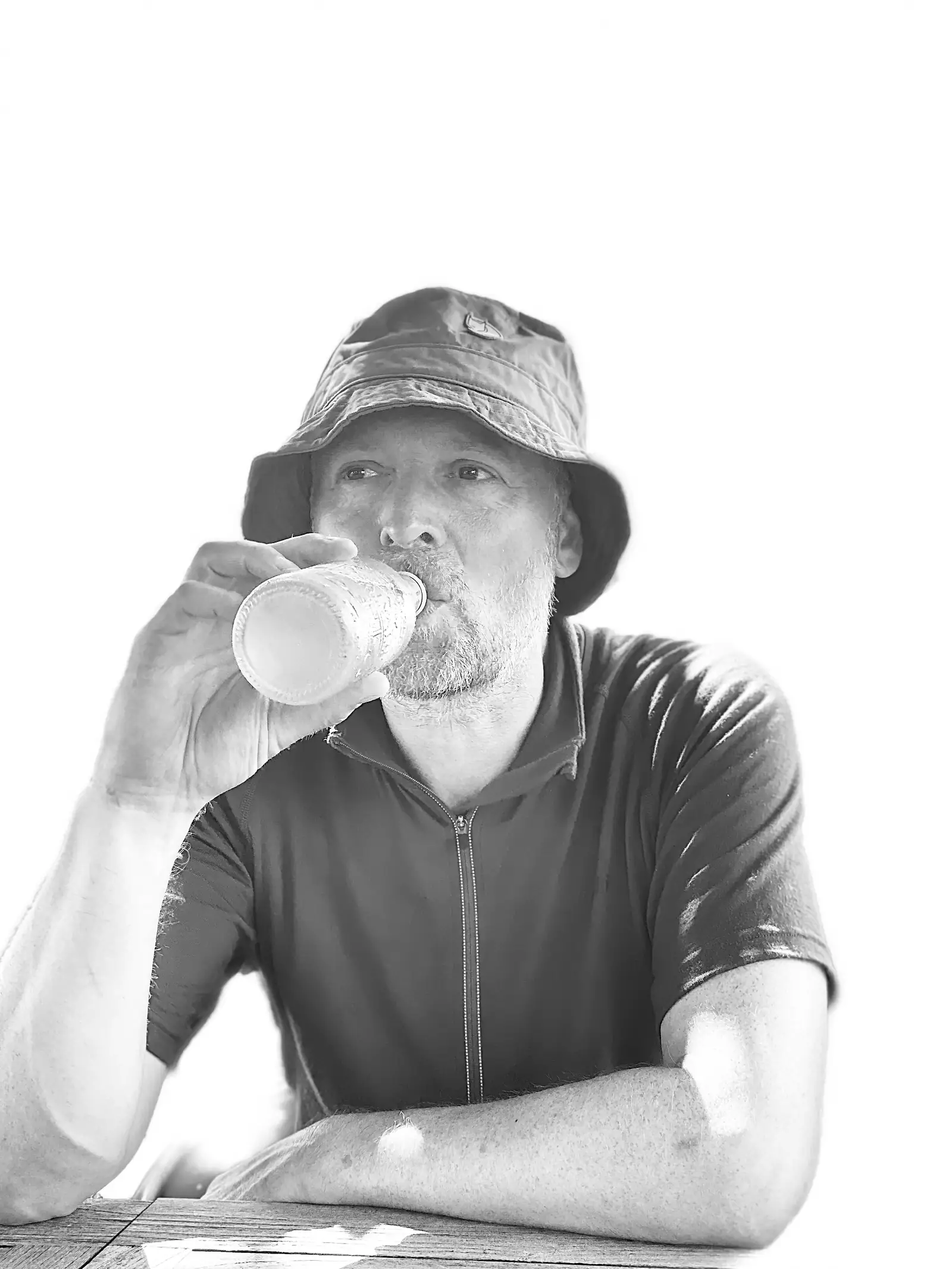 A bright high-key black-and-white portrait of a bearded person in a bucket hat drinking from a bottle at an outdoor table.
