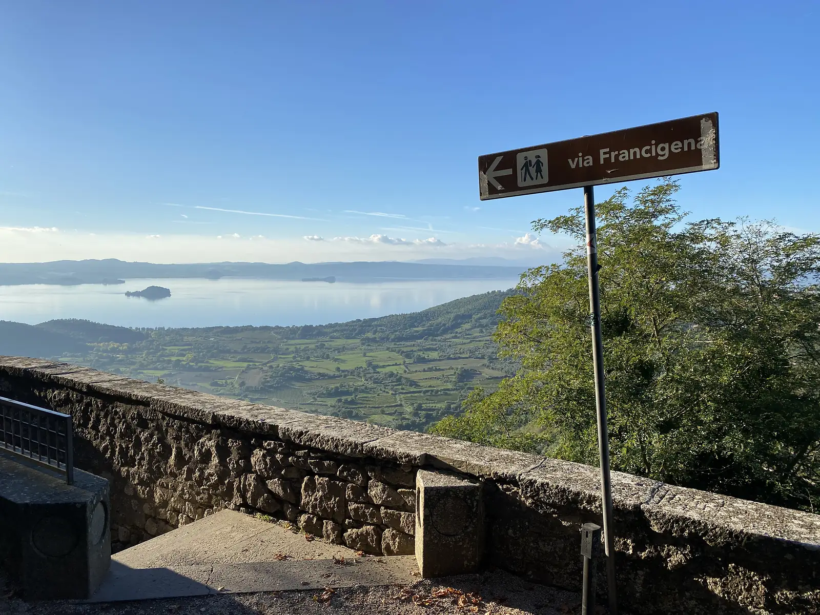 A "via Francigena" waymark points left from a stone parapet overlooking a wide lake dotted with small islands.