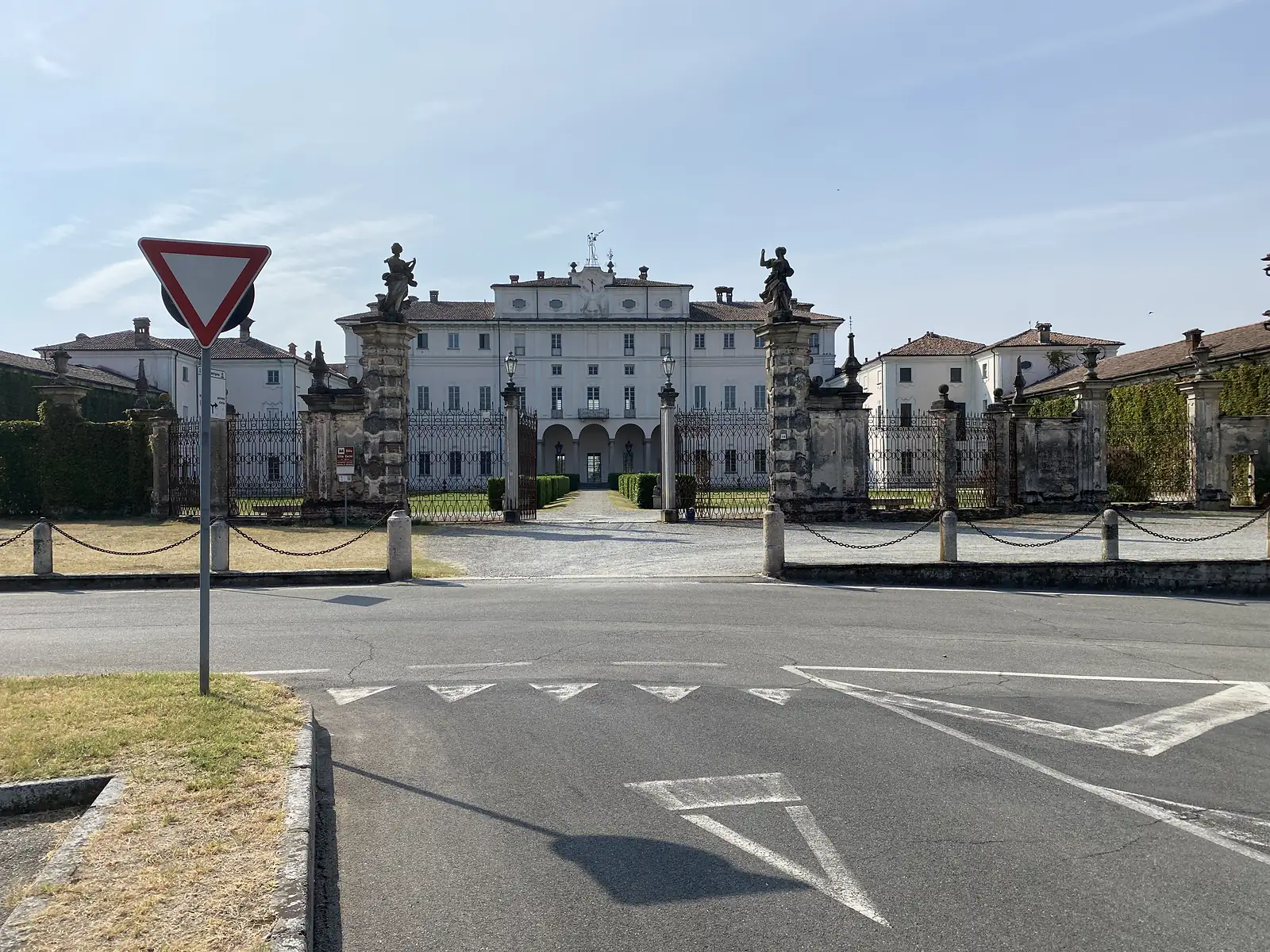 A give-way sign stands before the gated entrance to a grand white villa, its gate piers topped with statues, seen across an empty road junction.