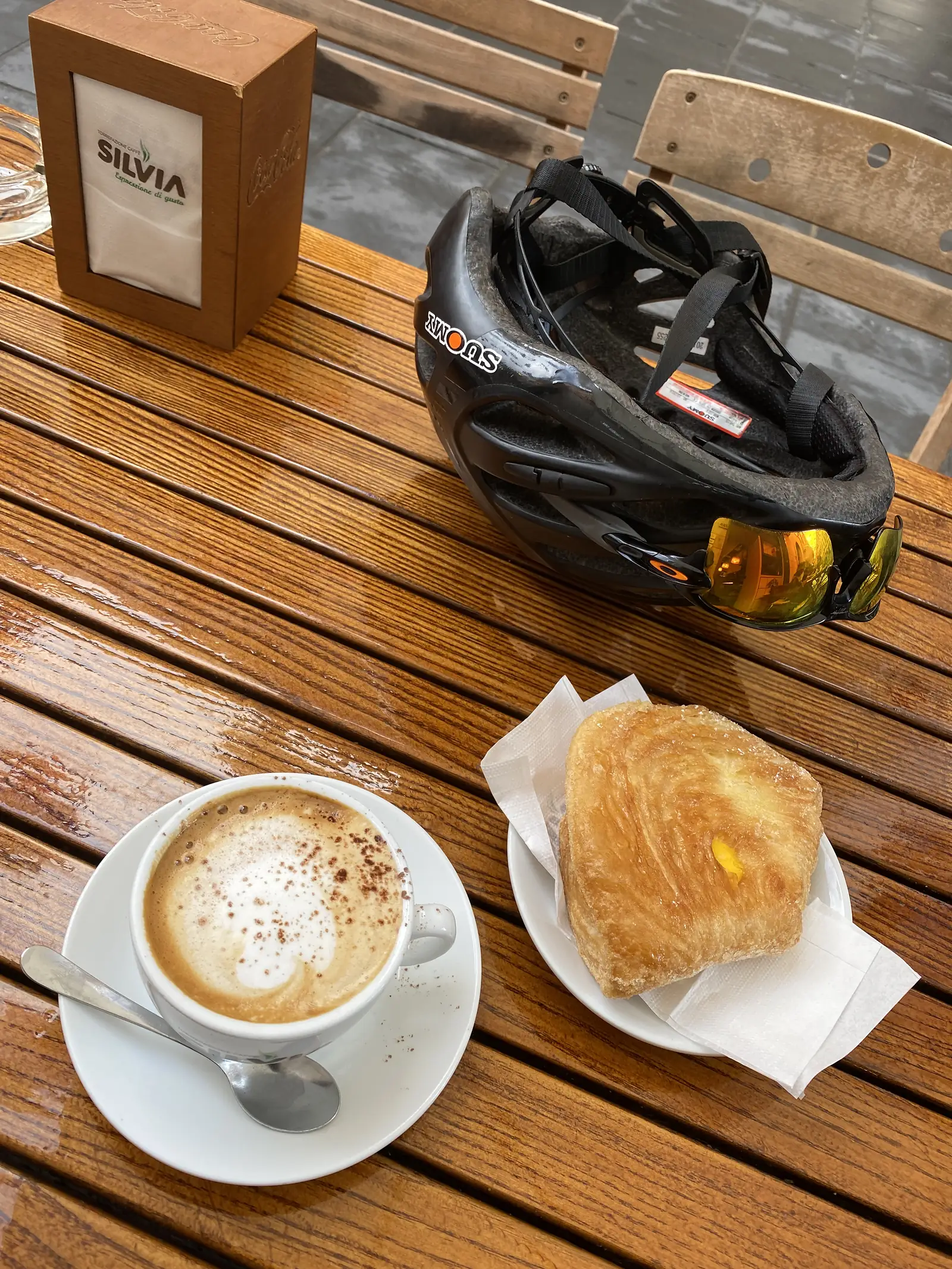A cycling helmet with sunglasses tucked into the vents sits on a café table next to a frothy cappuccino and a sugar-glazed pastry, seen from above.