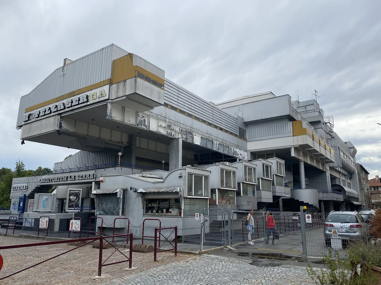 The derelict, streaked concrete facade of the former Hotel La Serra, its stacked guest-room pods cantilevered out above signs for a cinema and the Auditorium La Serra.