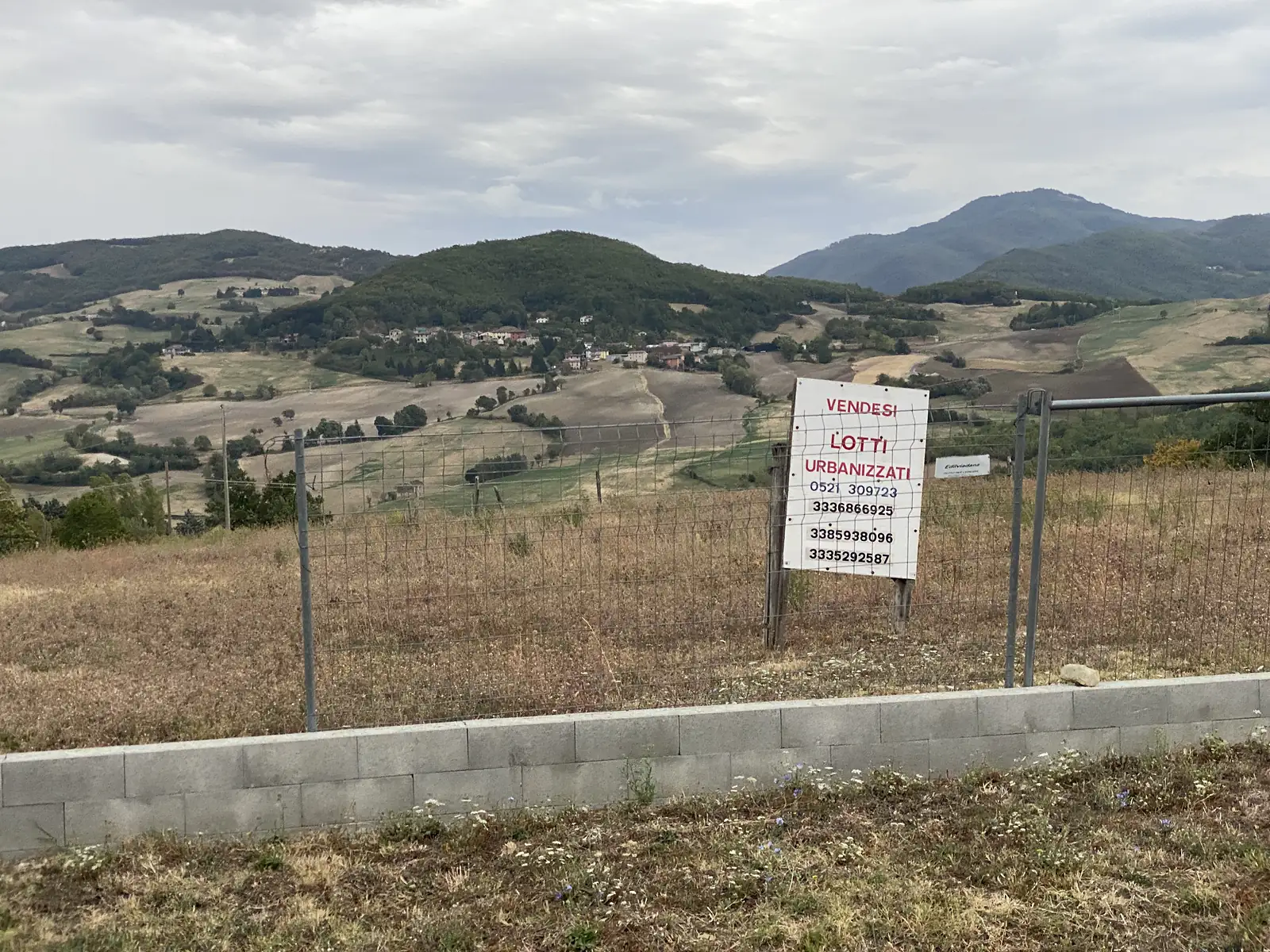 A hand-painted 'Vendesi lotti urbanizzati' estate agent's sign with phone numbers is wired to a fence overlooking a hamlet nestled among wooded Apennine hills.