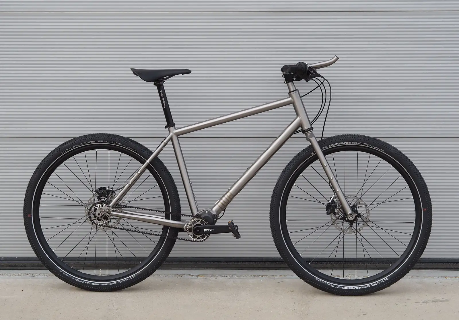 A flat-bar adventure bike side on against a corrugated steel shutter, its unpainted titanium frame built around a Pinion gearbox and a belt drive, on knobbly tyres.