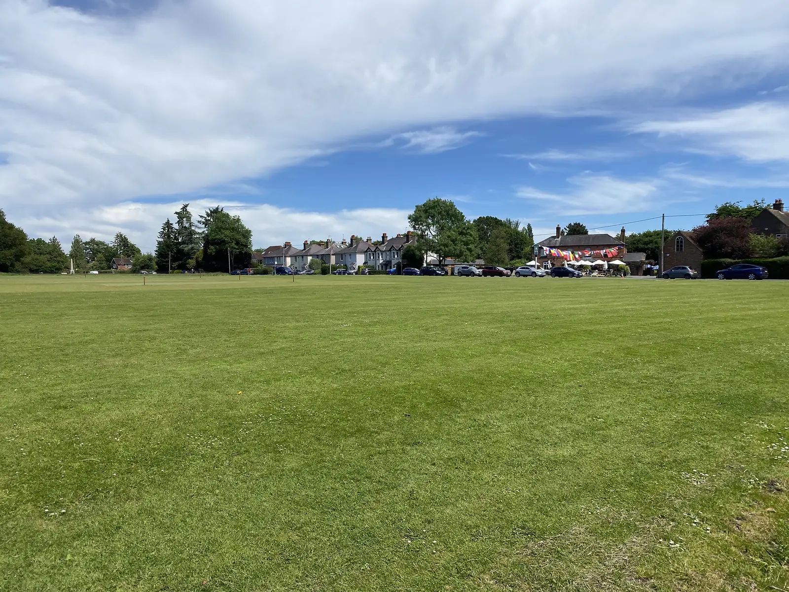 A wide mown village green with cricket stumps set up, houses and a bunting-strung pub along the far edge.