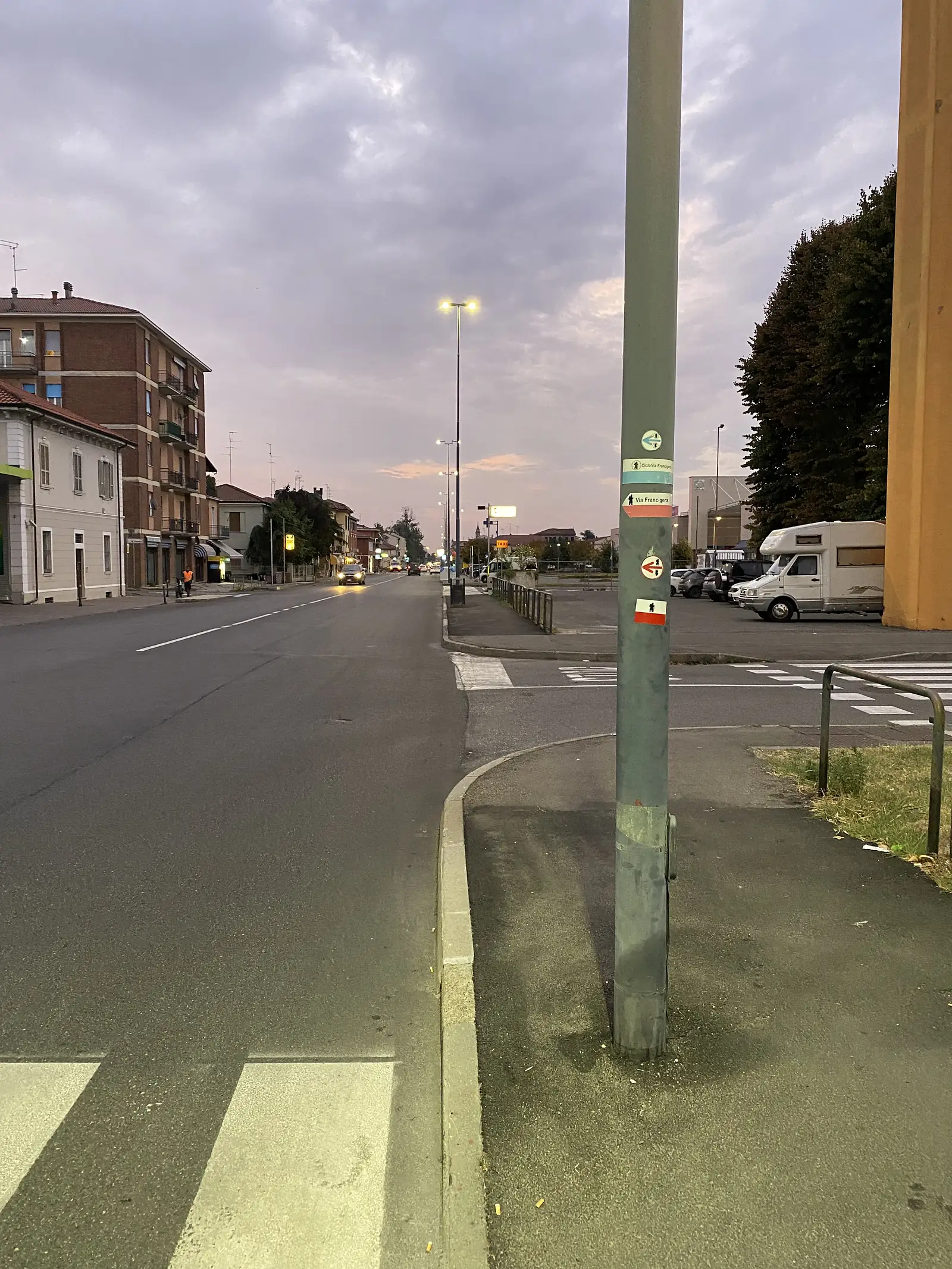 A green Via Francigena waymarker post on a town street at dusk, its pilgrim-and-arrow signs pointing the route past parked motorhomes and a row of streetlamps.