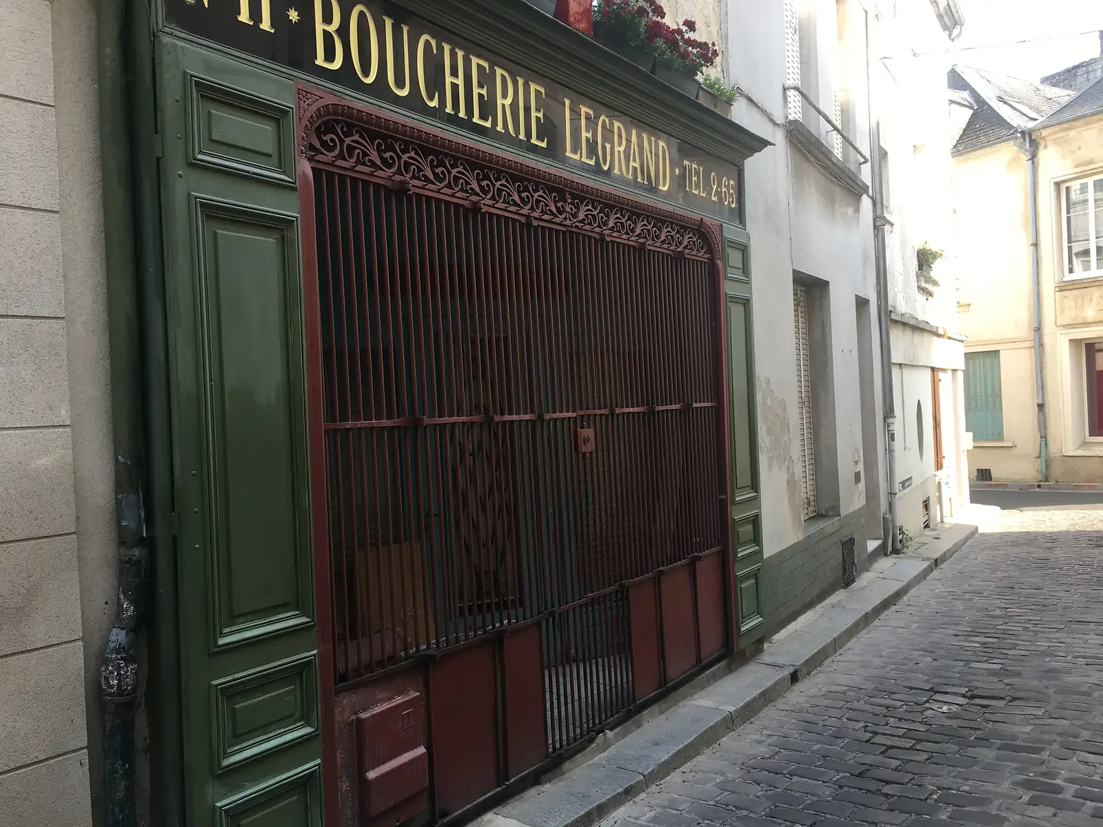 The shuttered shopfront of 'Boucherie Legrand' with gilded lettering and a decorative iron security grille stands on a sloping cobbled street.