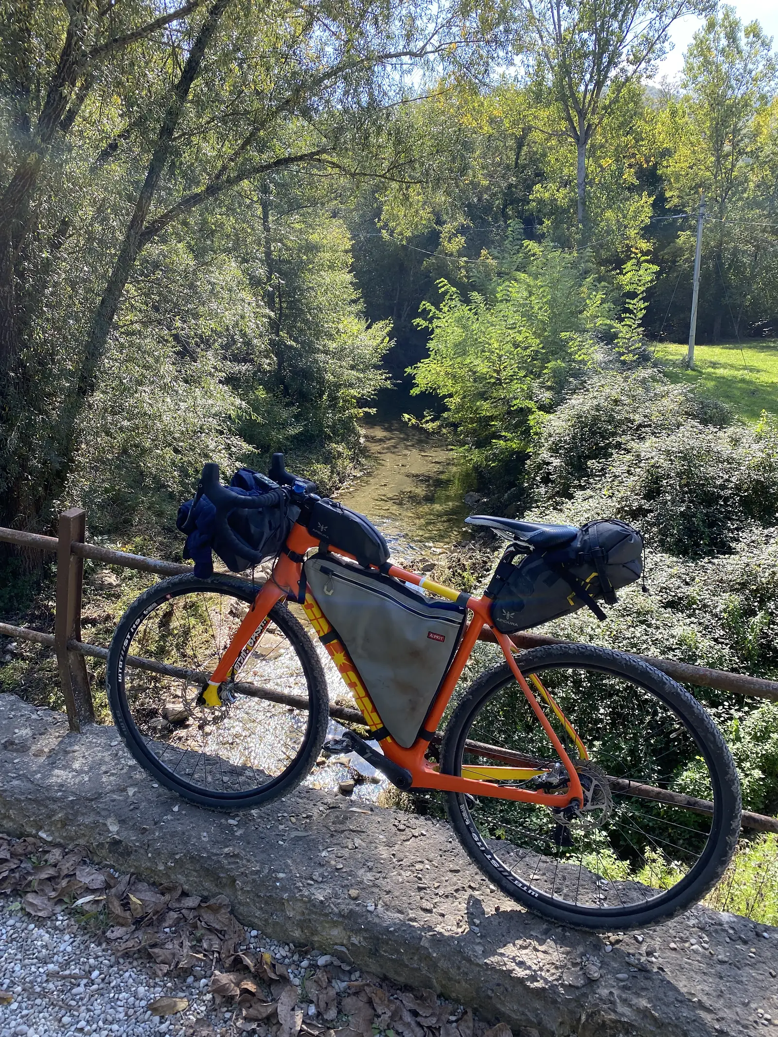 A loaded orange bikepacking bike propped against a low stone wall beside a calm, tree-lined stream.