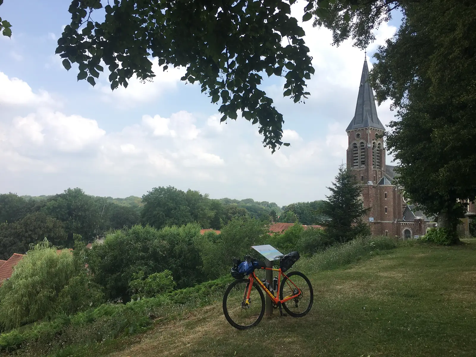 An orange loaded touring bike leaning against an information post on a grassy hilltop overlooking a village with a red-brick church tower and spire.
