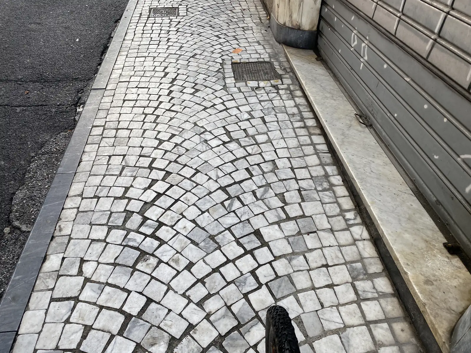 Close-up of a fan-patterned marble cobblestone pavement, worn smooth and pale grey-white, with a bicycle tyre entering the frame beside a marble kerb.