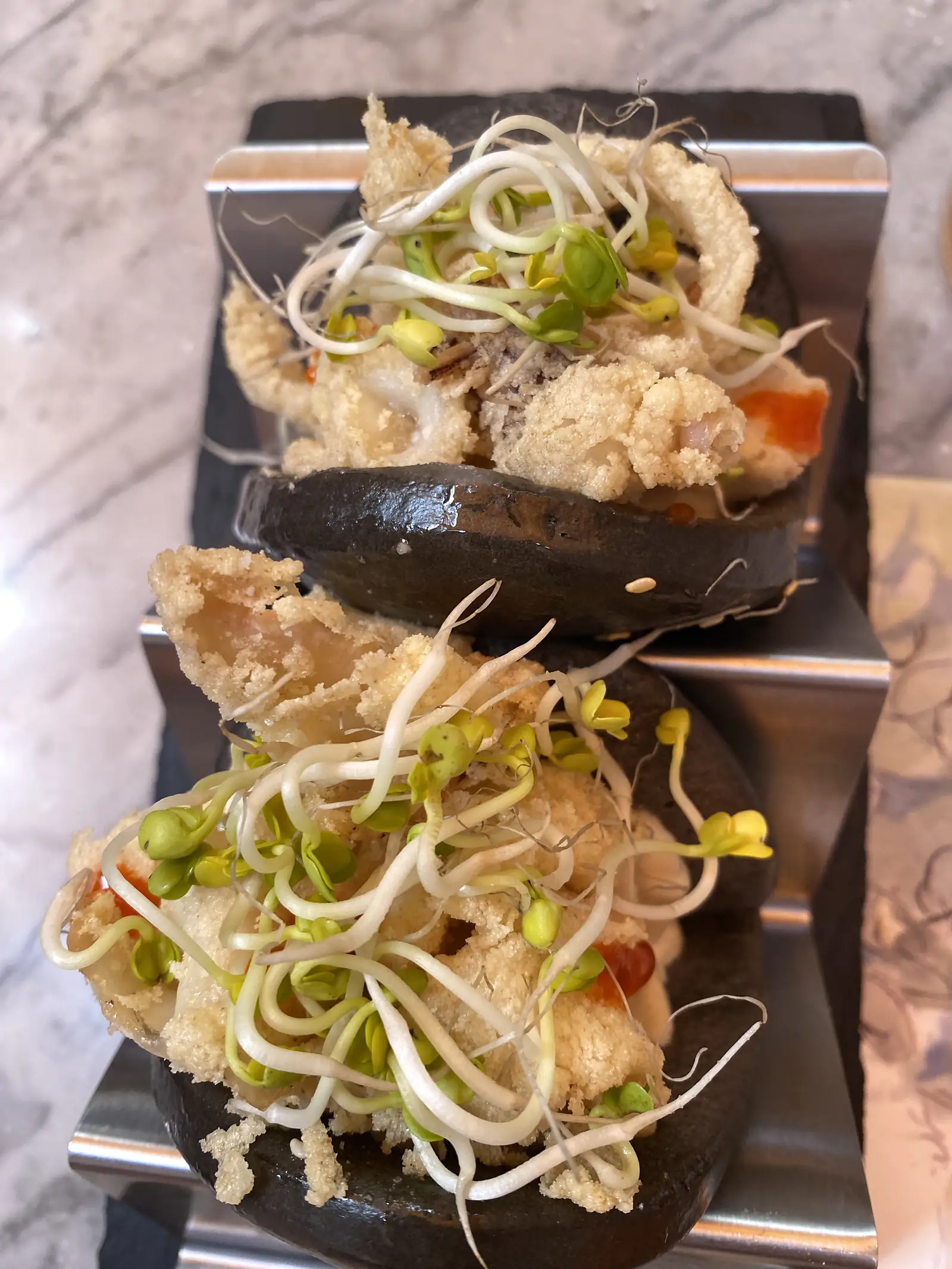 Two black buns filled with battered fried fish and topped with bean sprouts, served on a stepped steel stand.