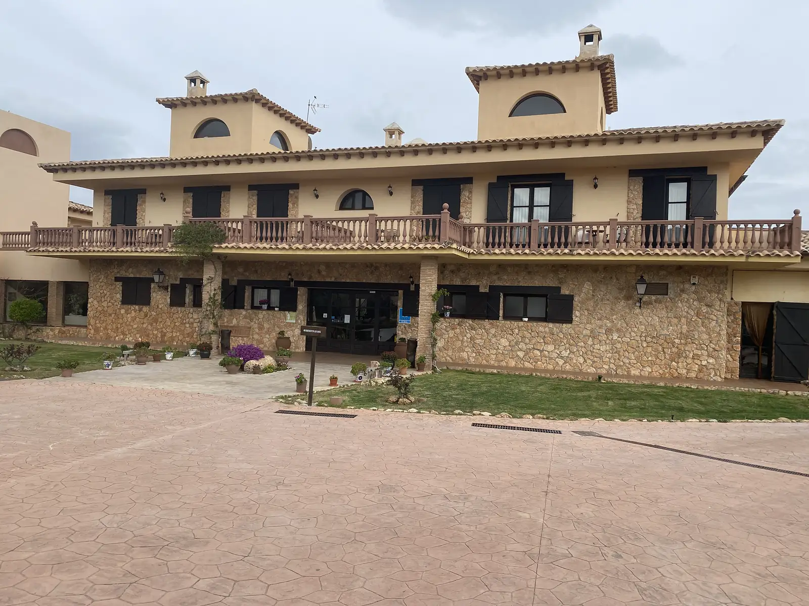 A two-storey country hotel of stone and cream render with a balustraded terrace and shuttered windows, on an empty paved forecourt.