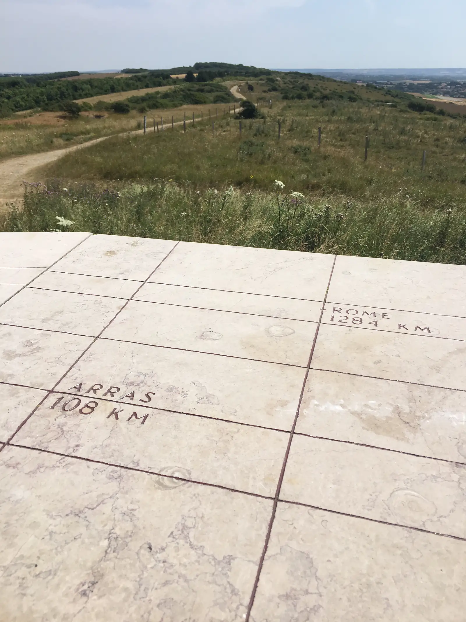 A stone orientation table engraved with distances to Rome (1284km) and Arras (108km), with grassland and the coast in the background.