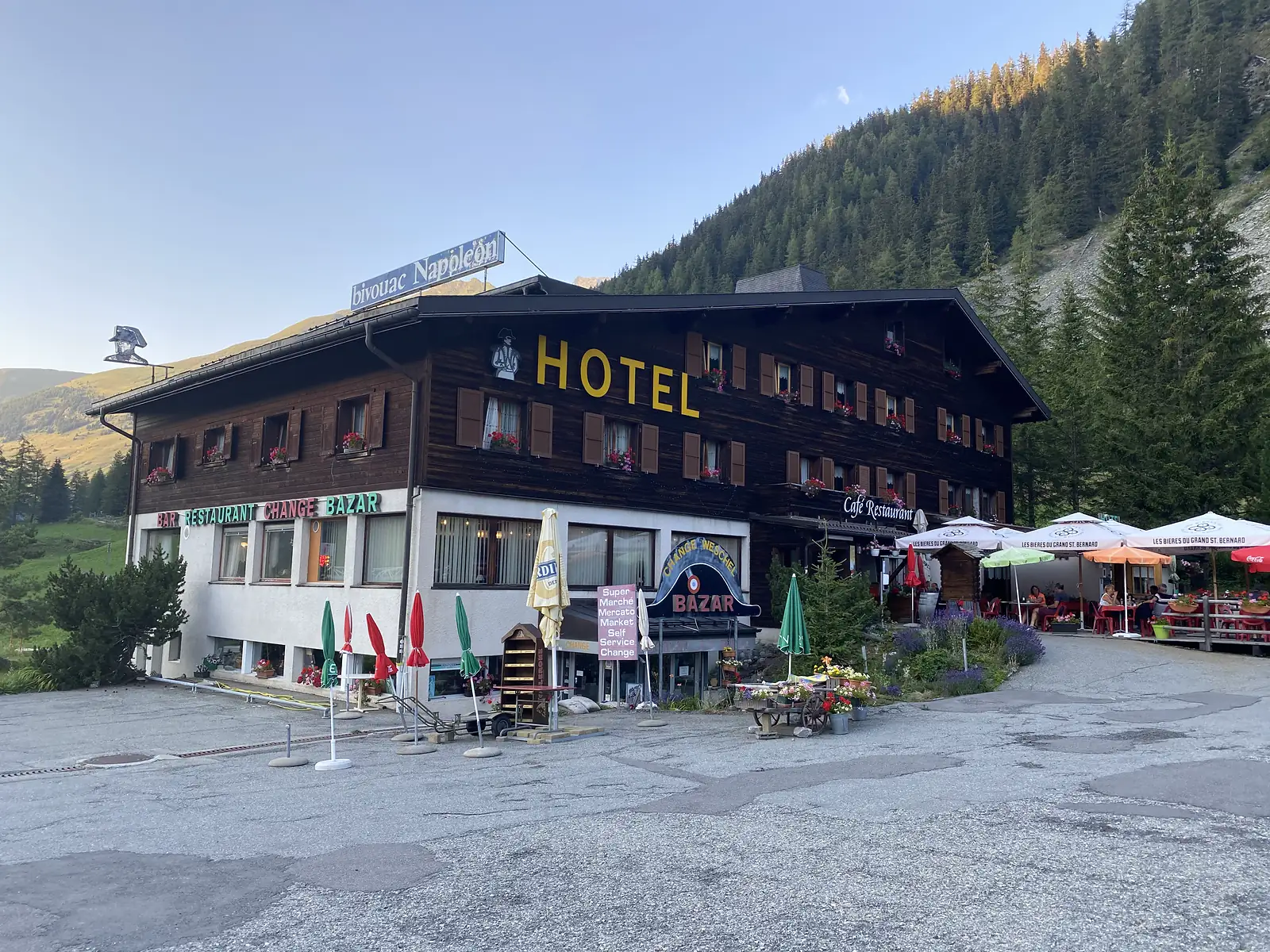 A dark-timbered mountain hotel signed Bivouac Napoléon and Hotel, its terrace crowded with parasols and diners at the foot of a forested slope.