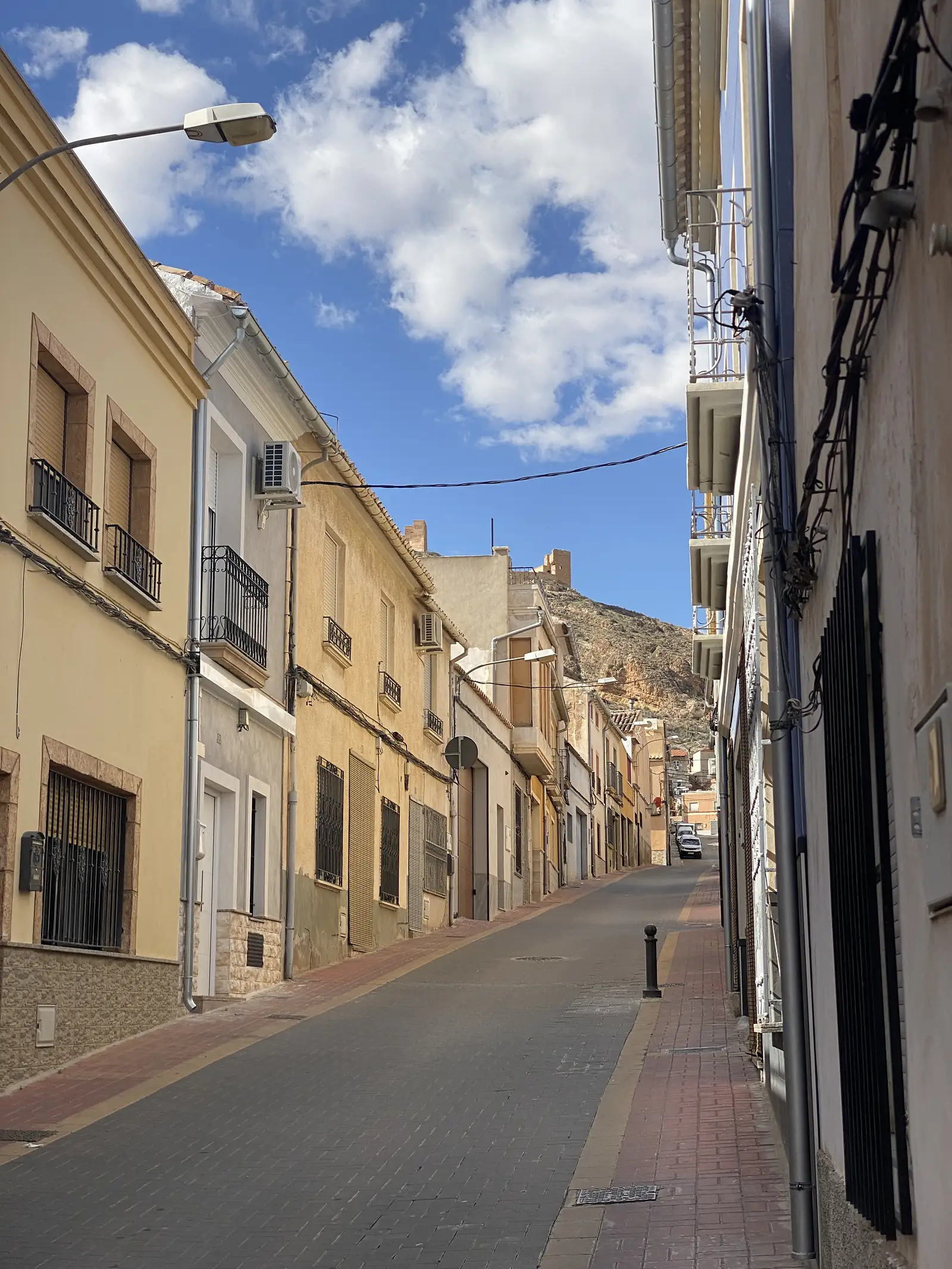 A quiet street of ochre houses with iron balconies looking towards a bare rocky hill topped by a ruined tower.