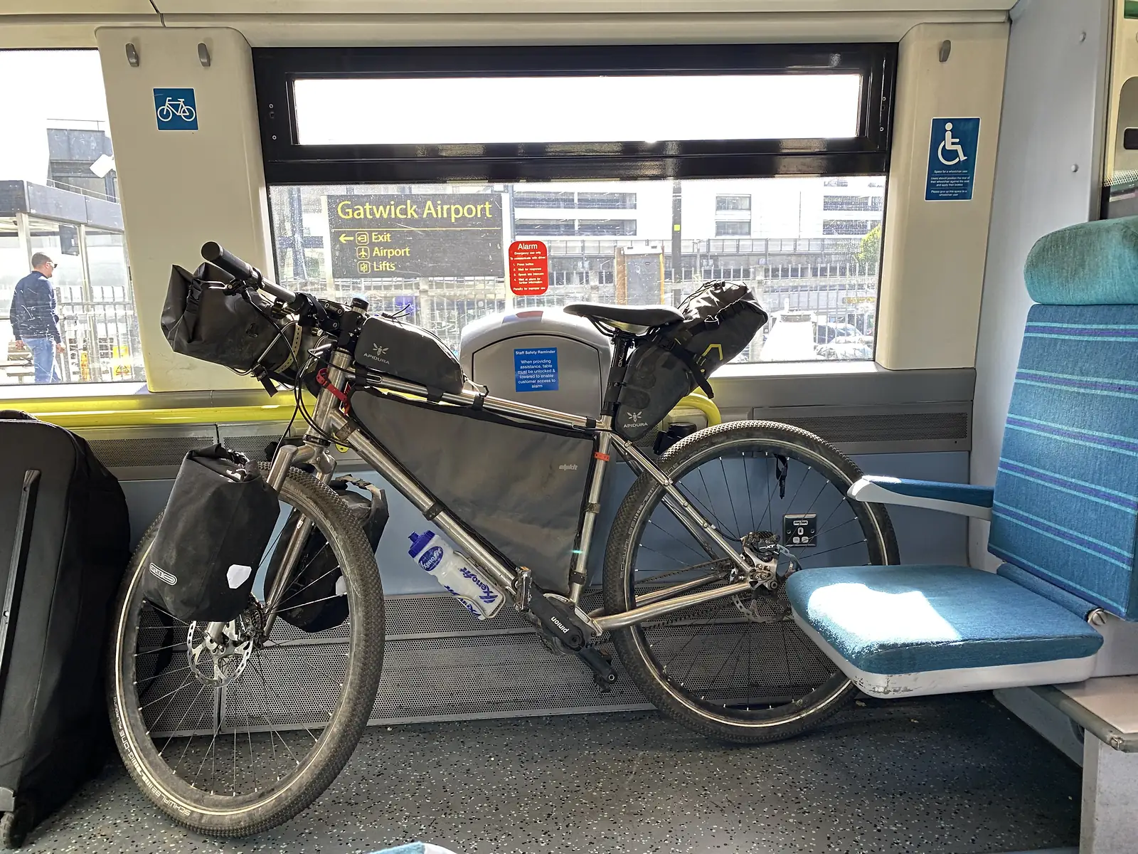 A loaded bikepacking bike standing in a train's cycle bay, with a platform sign reading "Gatwick Airport" through the window.