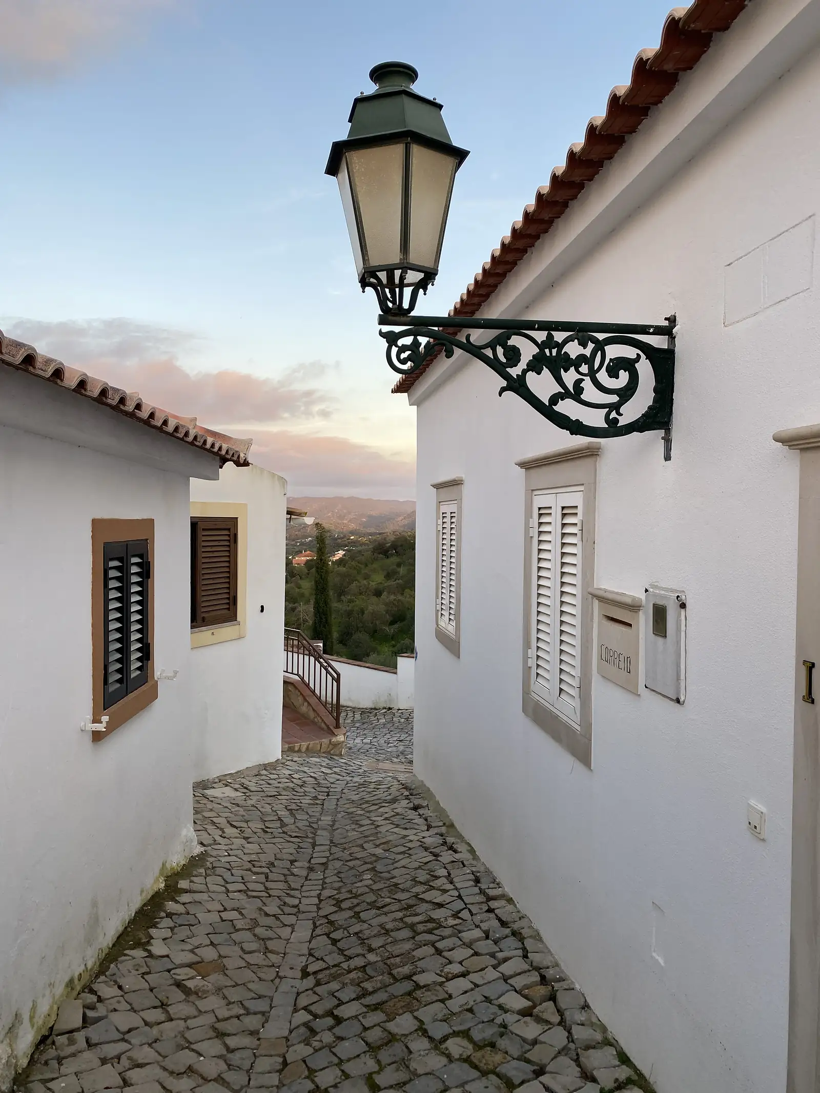 Ornate wrought-iron street lantern bracketed to a white wall above a narrow cobbled alley, which drops between shuttered houses towards hills under a pink dusk sky.