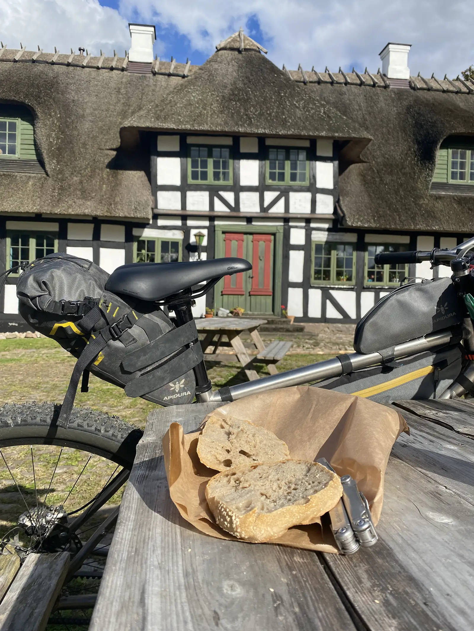 A halved sourdough loaf on brown paper on a picnic table beside a loaded bike, with a thatched half-timbered house behind.