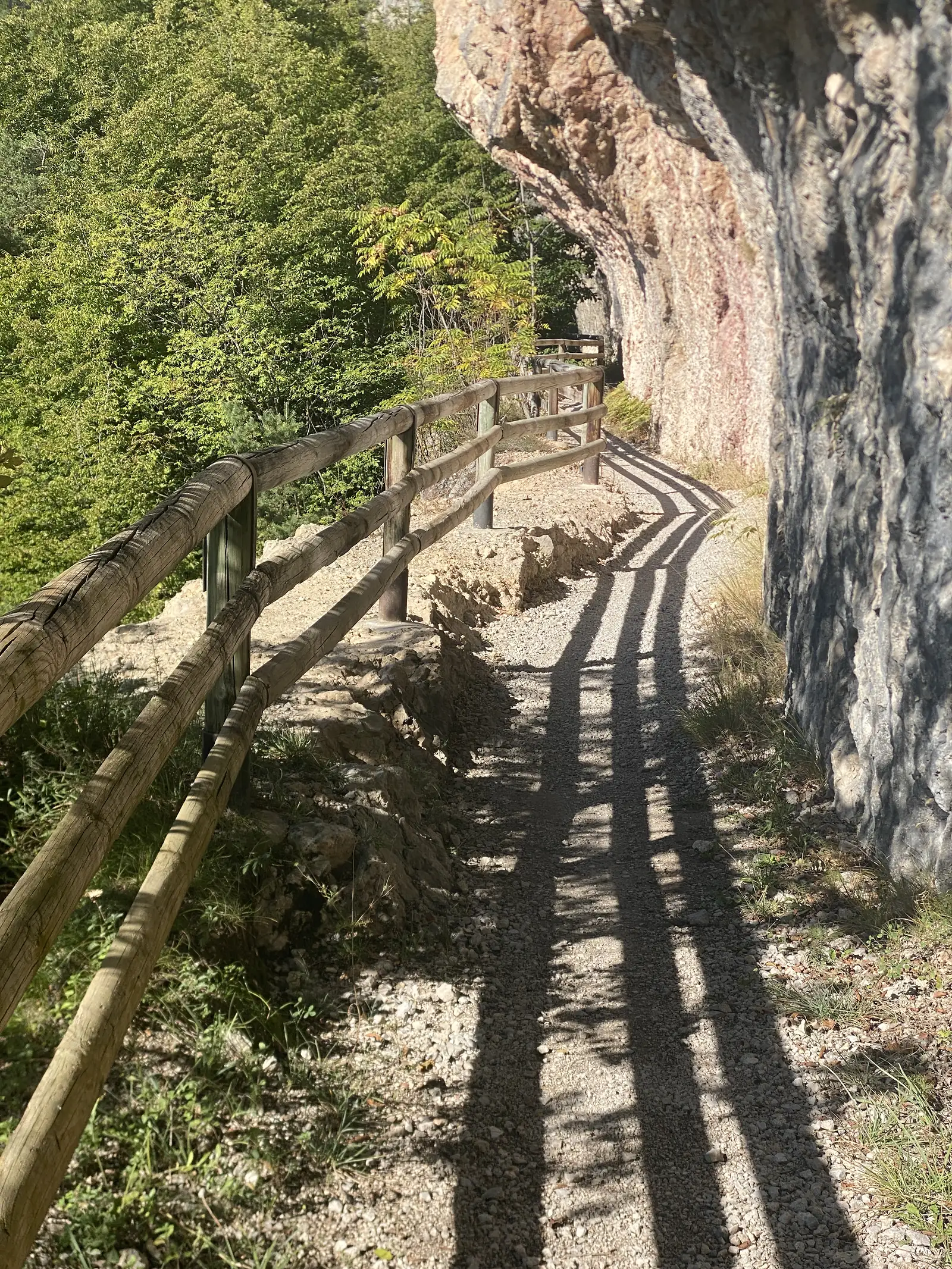 The gravel path squeezes beneath an overhanging limestone cliff, a timber fence on the outside edge and fence shadows striping the surface.