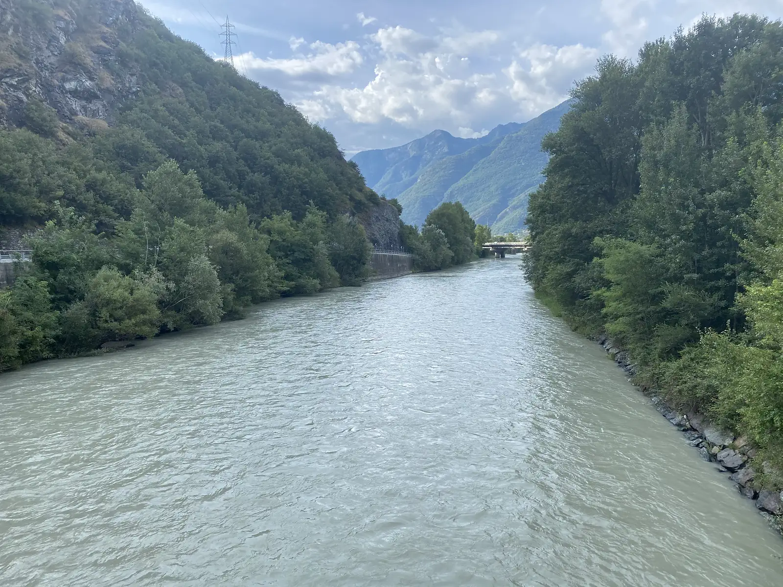 A wide grey-green river flowing between wooded banks towards distant mountains, with a road bridge crossing it further downstream.