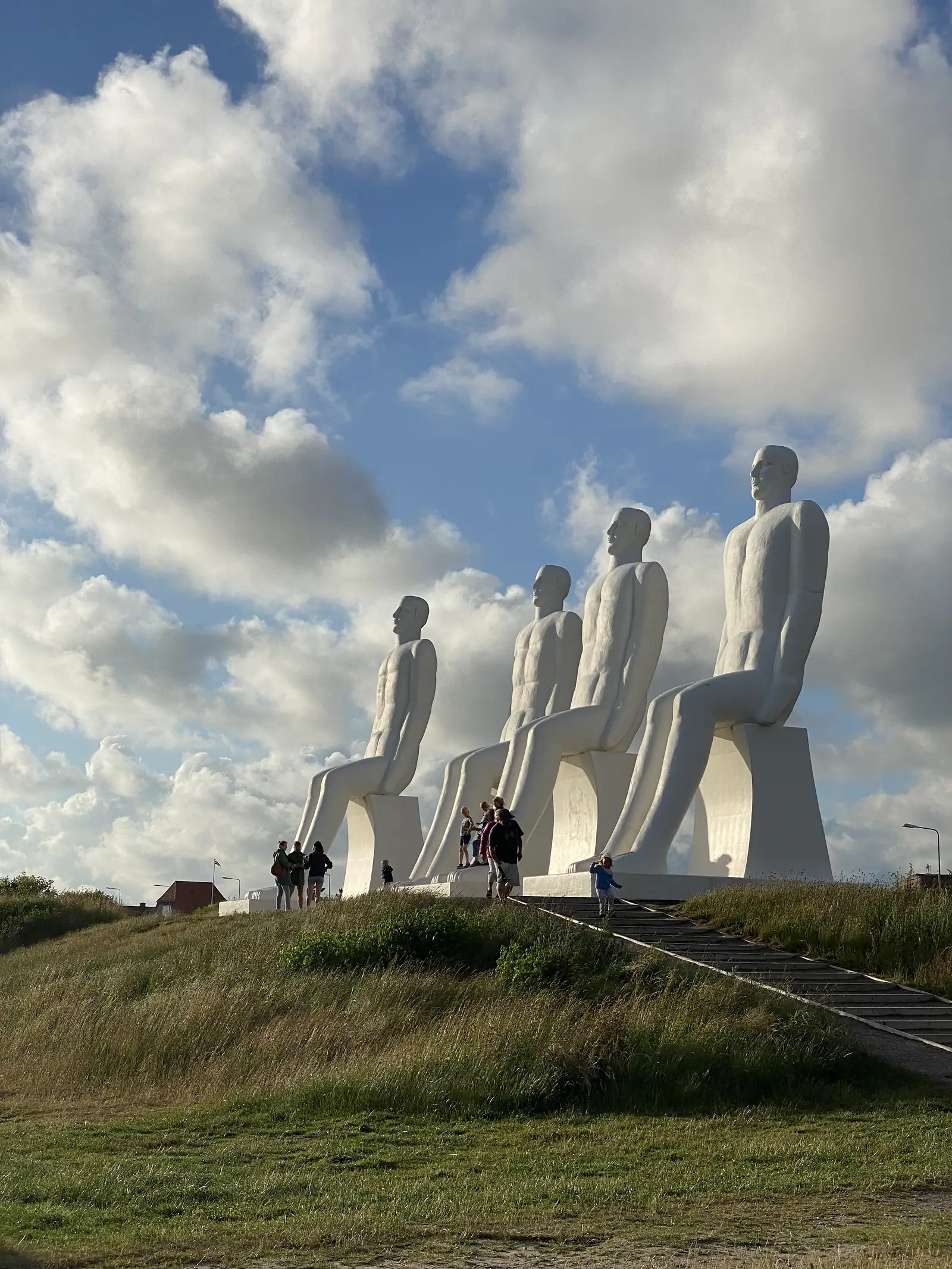 Four colossal white seated figures on a grassy rise above a flight of steps, with people dwarfed at their feet under a towering cumulus sky.